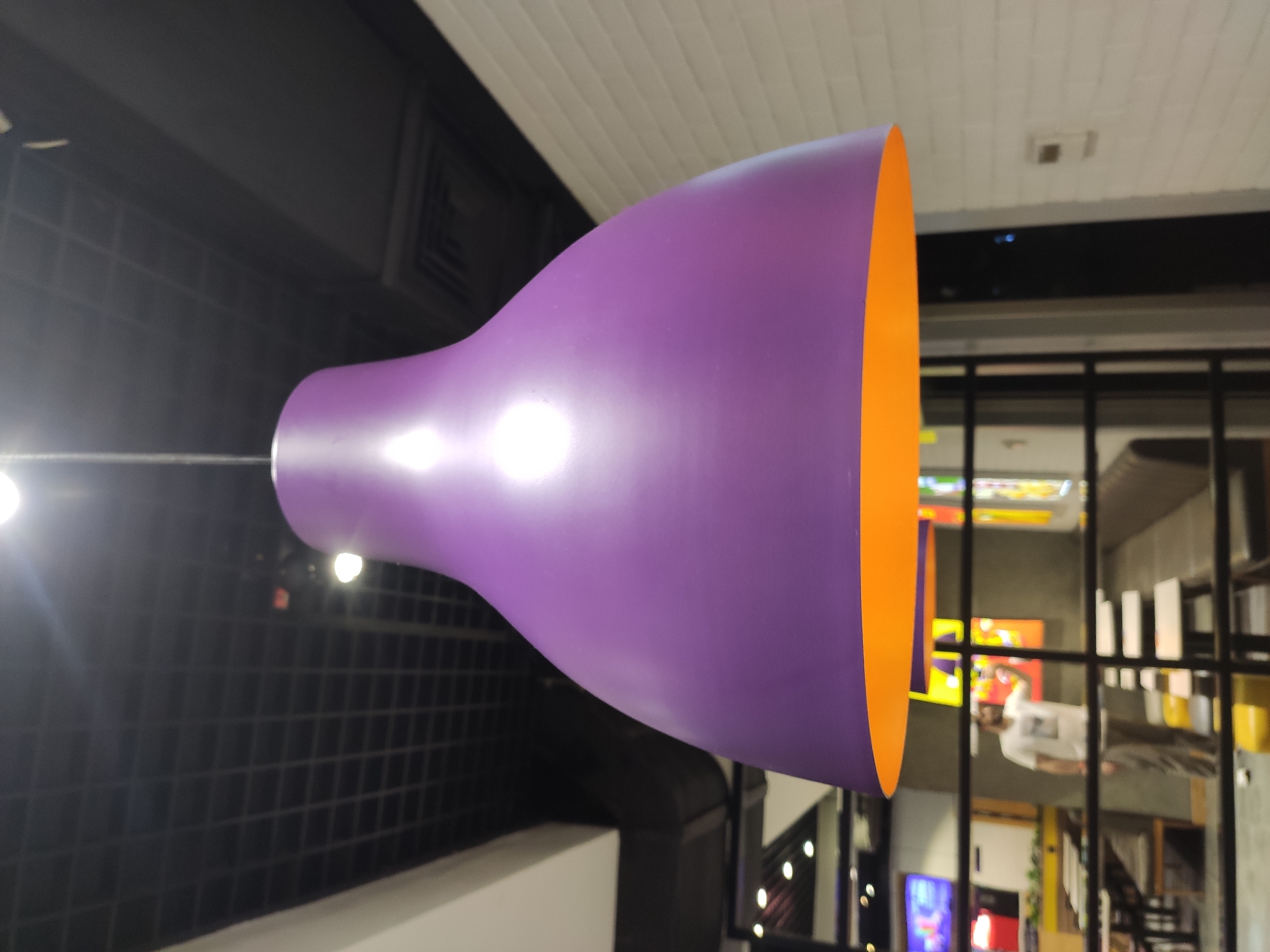 A purple bell-shaped pendant light with an orange interior hangs from the ceiling. The ceiling is made of black acoustic tiles. In the background, a man in a white t-shirt and grey pants is standing and holding a phone, possibly taking a selfie. The man is blurred and appears to be in a restaurant or fast-food establishment, with tables and seating visible. The environment is indoors, with some lighting and possibly screens on the wall. The location is identified as Tashkent, Uzbekistan.