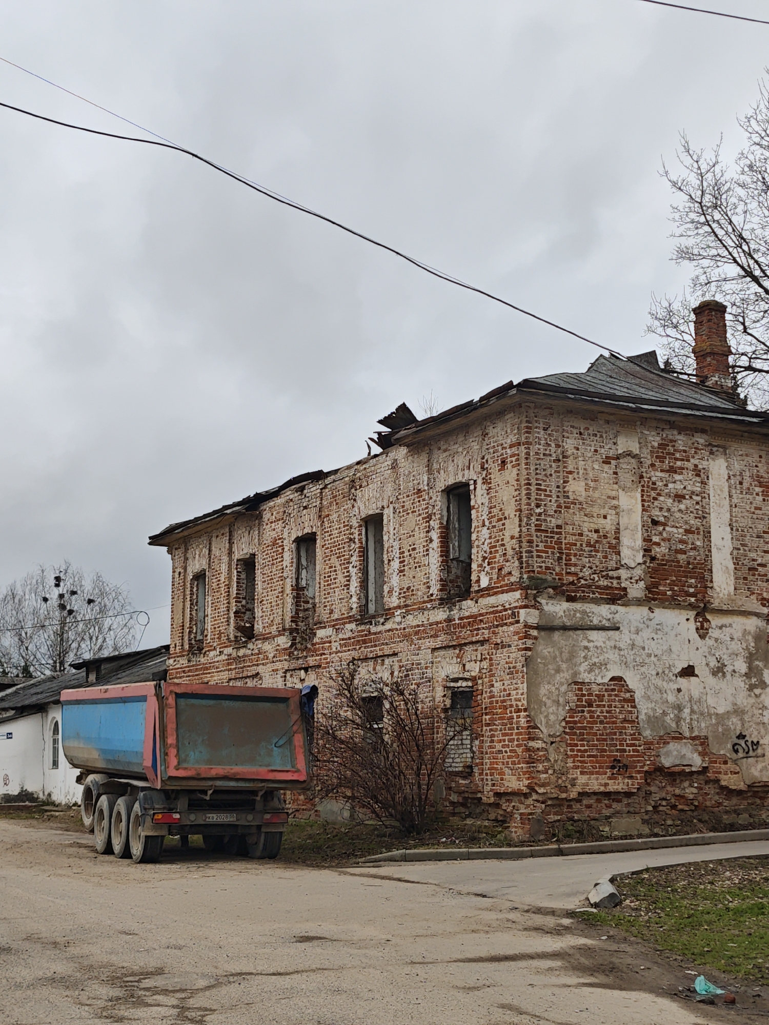The image depicts a dilapidated two-story brick building with missing windows and crumbling facade, located in посёлок совхоза "Буденновец", Russia. A dump truck with a blue and red bed is parked on a dirt road in front of the building. The sky is overcast. A few bare trees are visible. A white fence is partially seen on the left. A power line stretches across the top portion of the image. Graffiti is visible on the lower portion of the building's right side. The building appears abandoned and in disrepair.