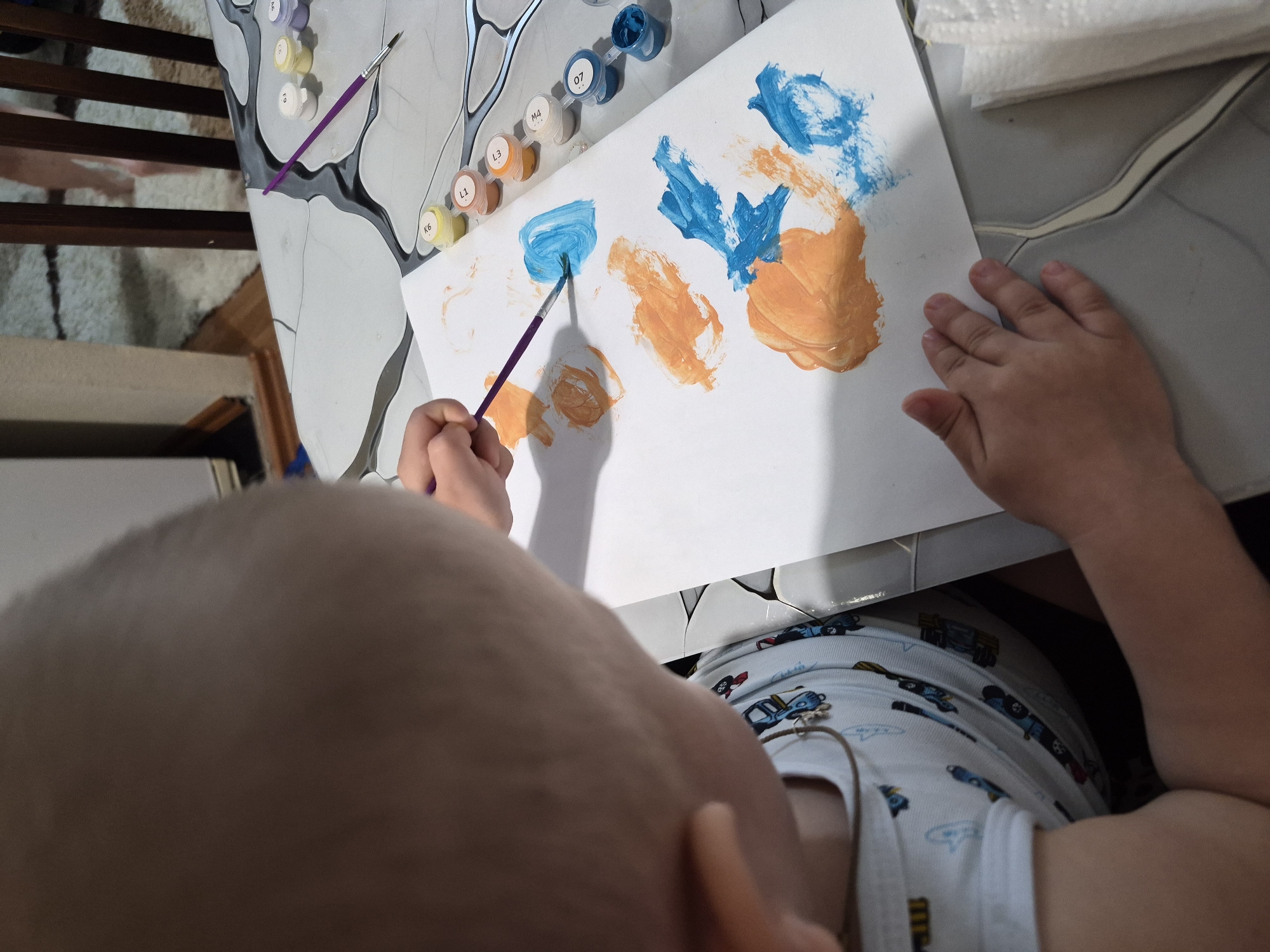 A child is painting on a piece of paper at a table. The child is using a paintbrush and paint-by-numbers kit. The child is wearing a white shirt with a car pattern. The table is gray and white and is next to a wooden staircase. The table is in a room that appears to be a living space. The location is not evident, and there is nothing that identifies the location as Tobolsk, Russia.