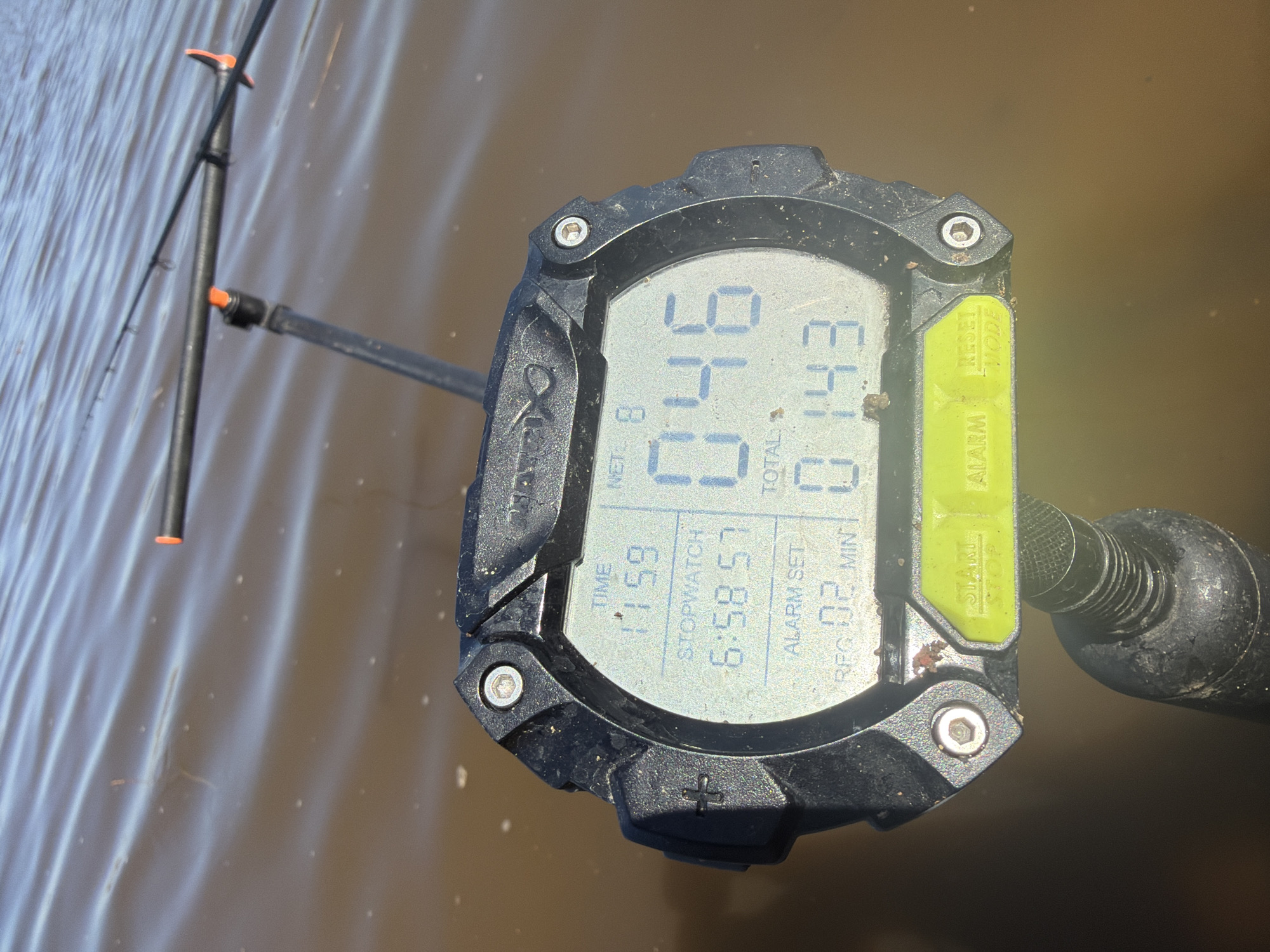 The image shows a waterproof watch partially submerged in water. The watch face displays digital information, including "TIME 11:59," "STOPWATCH 6:58.57," "NET 8," "TOTAL 0:14.3," and "RPG 02 MIN." The watch has physical buttons labeled "ALARM," "START STOP," "RESET MODE," and a button with a "+" symbol. A portion of a fishing rod or similar pole is visible in the background of the image on the left side of the watch. The water appears somewhat murky, as if it is natural water, not from a swimming pool.