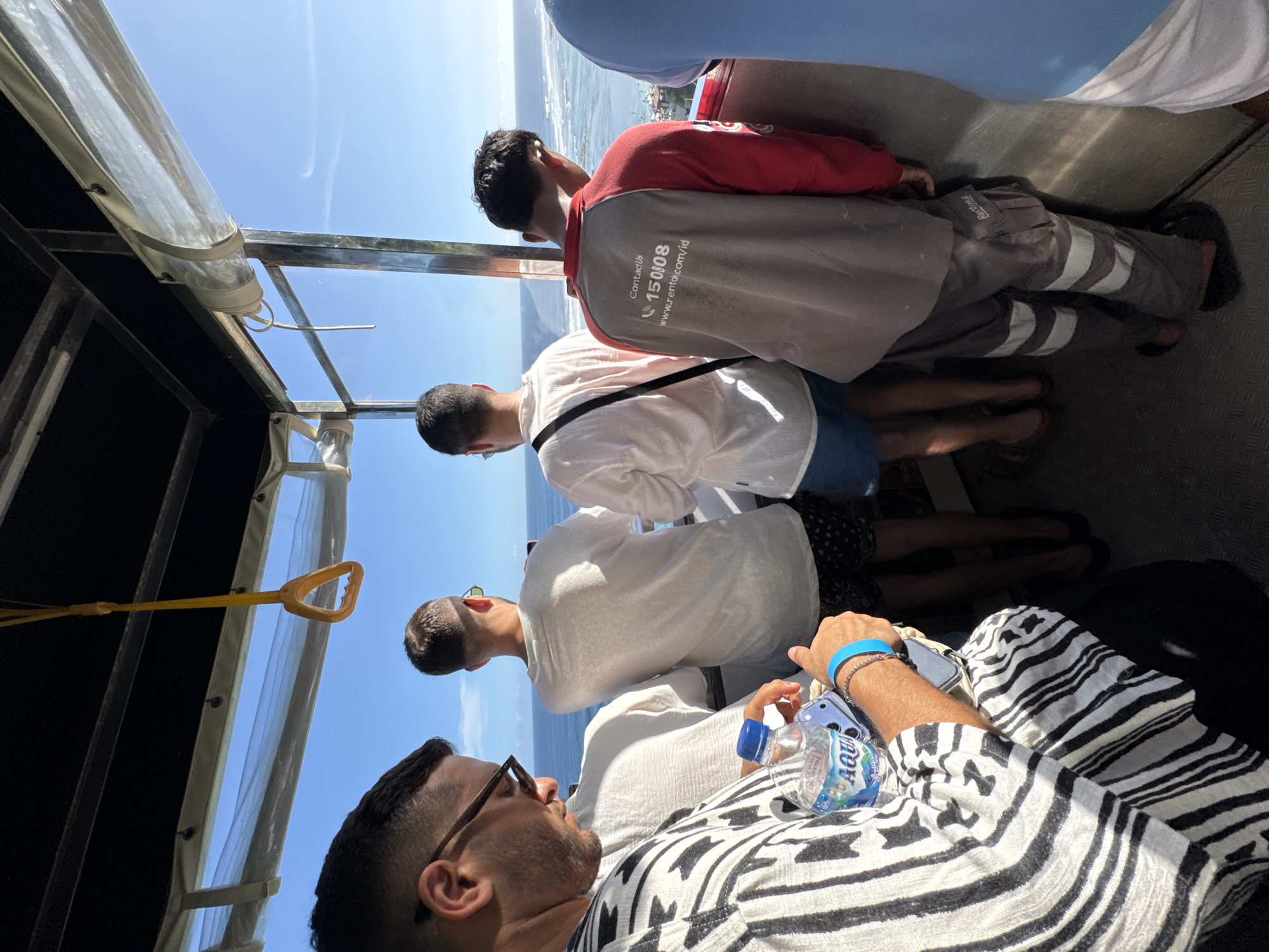 Here's a description of the image:
The image captures a group of people inside a vehicle, possibly a boat or bus, looking out toward the sea. The location appears to be Kuta Selatan, Indonesia, as indicated by the contact details "Contact us: 150808 www.rental.com/id" on the red shirt of the person closest to the sea.
Several individuals are visible in the frame, mostly from the back or side. One man in the foreground wears a black and white patterned shirt, glasses, and has a water bottle and a smartphone next to him. Further back, there are three men wearing white shirts and casual bottoms, their heads turned toward the view. Another man in a red shirt with grey pants is standing next to them looking at the sea.
The vehicle has a partially transparent roof, allowing the sunlight to illuminate the interior. A yellow handle is suspended from the roof. The sea is visible in the background, with a hint of the coastline.