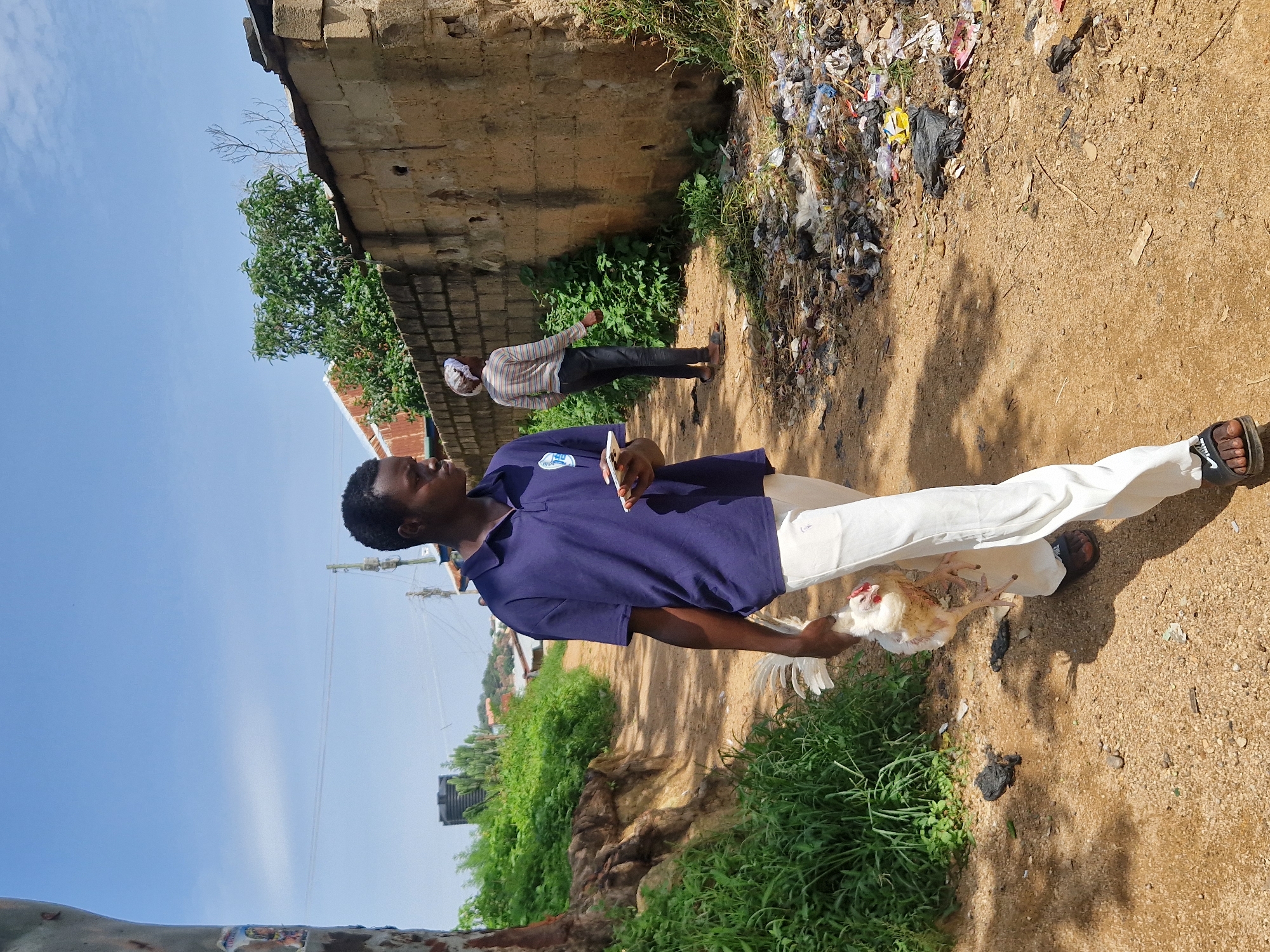 A young Black man is walking on a dirt path, holding a white chicken in his left arm. He is wearing a dark blue polo shirt and light-colored pants, with dark sandals on his feet. In his right hand, he is holding a white smartphone. He is looking to his right. In the background, a woman in a striped shirt and headscarf is walking away from the camera. To the right of the path is a rough, stone wall piled with garbage. To the left of the path are green plants and a tree trunk. The sky is blue with some clouds.