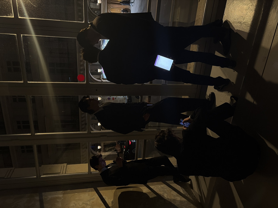 The image depicts five adult males, all dressed in dark formal attire, positioned within a dimly lit interior space looking out through a large, multi-pane window onto a city street at night. The architectural style visible outside, including characteristic buildings, streetlights, and a red circular traffic signal, indicates the location is Paris, France.
In the foreground, one male sits on the floor, leaning forward, engaged with a smartphone that emits a faint blue light. To the left, a standing male leans against the window frame, looking down at his own smartphone. Centrally, another male stands upright, looking towards the right. On the far right, two males stand close together; one is holding an illuminated tablet or smartphone, its bright screen visible and casting light onto their lower torsos. All individuals are largely silhouetted against the exterior light, obscuring facial details. The exterior scene reveals classical Parisian buildings, street-level activity with faint pedestrian figures, and vehicle lights. The interior wall to the left appears textured, possibly stucco or plaster.
