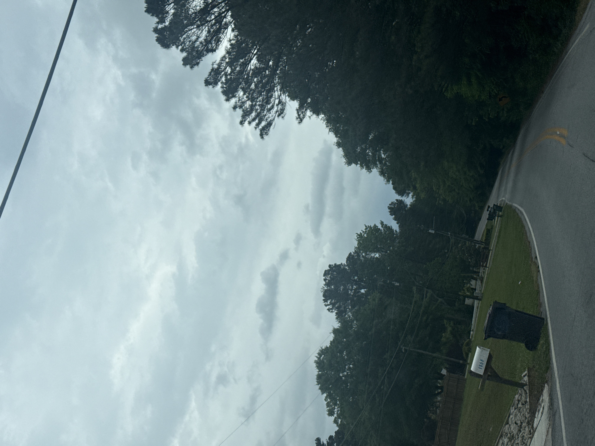 The image shows an overcast sky above a residential street in Mountain Park, United States of America. Power lines are visible against the sky. The street curves, bordered by trees and green lawns. A mailbox with a visible address of "1866" stands near the roadside. The road surface appears wet, possibly from rain.