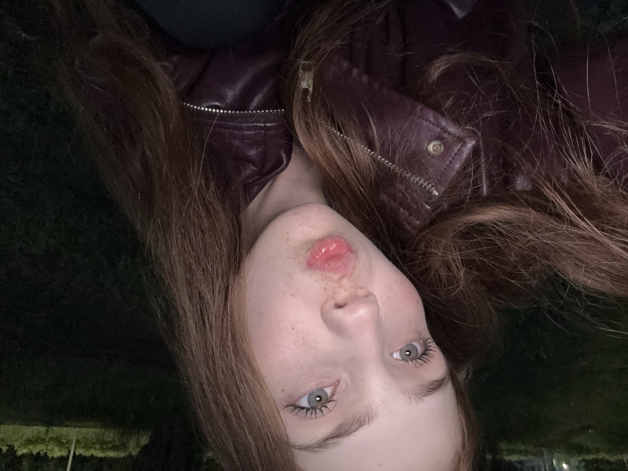 A young individual, appearing female, is depicted in a close-up, likely self-portrait, with long, reddish-brown hair cascading around the face and shoulders. The subject has light-colored eyes (appearing blue-green/hazel), with visible mascara on the upper lashes, and freckles scattered across the nose and cheeks. The lips are puckered in a "kissing" or "duck face" pose, enhanced by a glossy pink product.
The individual is wearing a dark, possibly maroon or burgundy, leather-style jacket. Details visible include silver zippers on the front and a silver snap button near the collar or lapel area.
The background is dark, heavily blurred, and out of focus, indicating an outdoor nighttime or very dimly lit environment. Indistinct dark green shapes, consistent with foliage or grass, are faintly visible, particularly in the upper left and right portions of the frame. Subtle, blurred light sources are discernible in the distant background, suggesting artificial illumination at an unspecified distance. No specific architectural landmarks, unique geographical features, or other identifying elements are present within the image to determine a precise location, including the city of Naberezhnye Chelny, Russia.