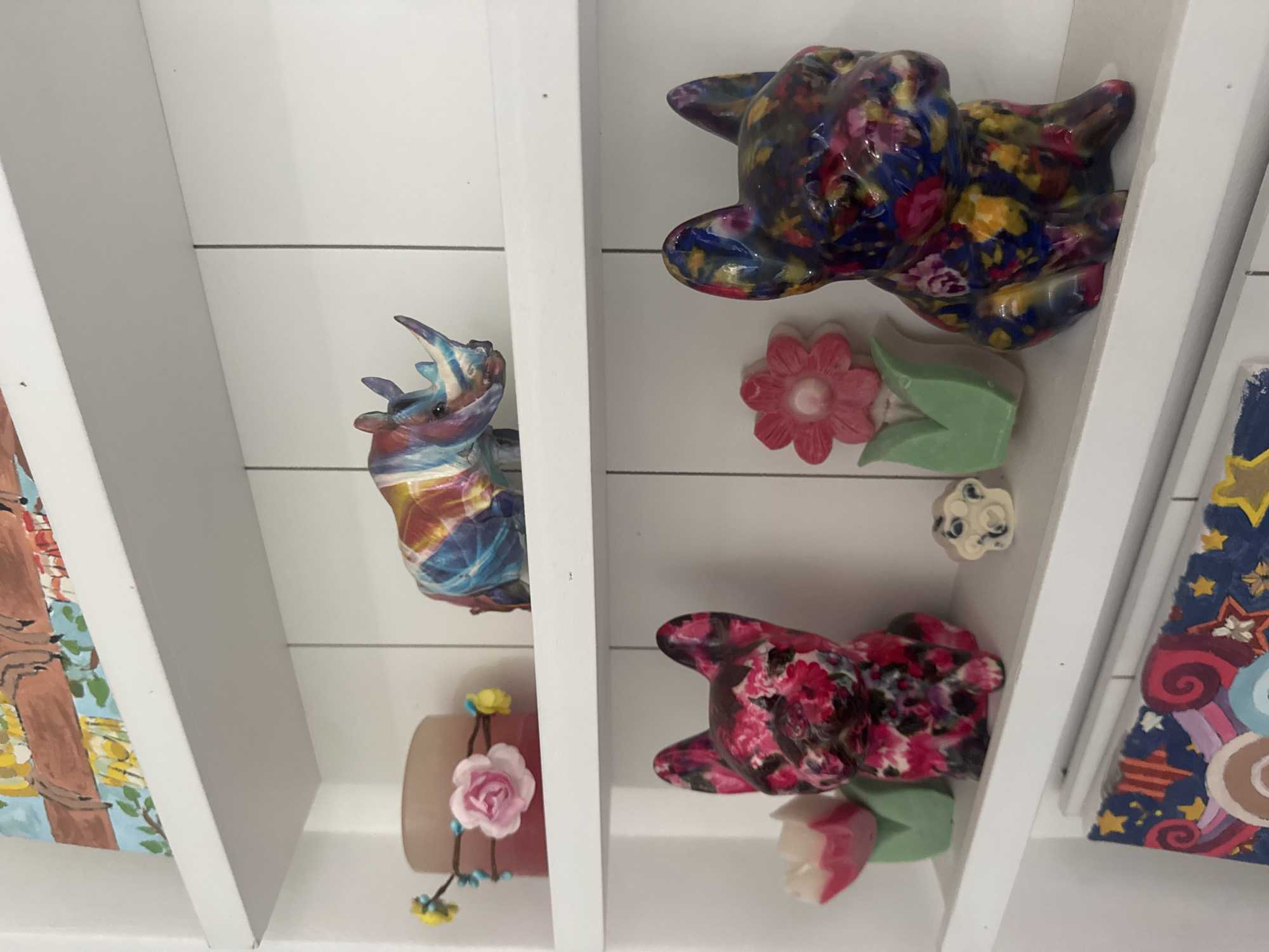 A low-angle shot displays a white shelf with horizontal planks holding several objects. On the left, there is a painting, followed by a colorful rhino figurine and a small decorative pot with a pink flower. In the next section, there's a floral-patterned dog figurine, pink flower and green tulip decorations, and a small white decorative item. On the right side of the shelf, there is another floral-patterned dog figurine and a painting. The overall impression is a curated shelf display. No people are visible. There is no information about Plovdiv, Bulgaria in the image.