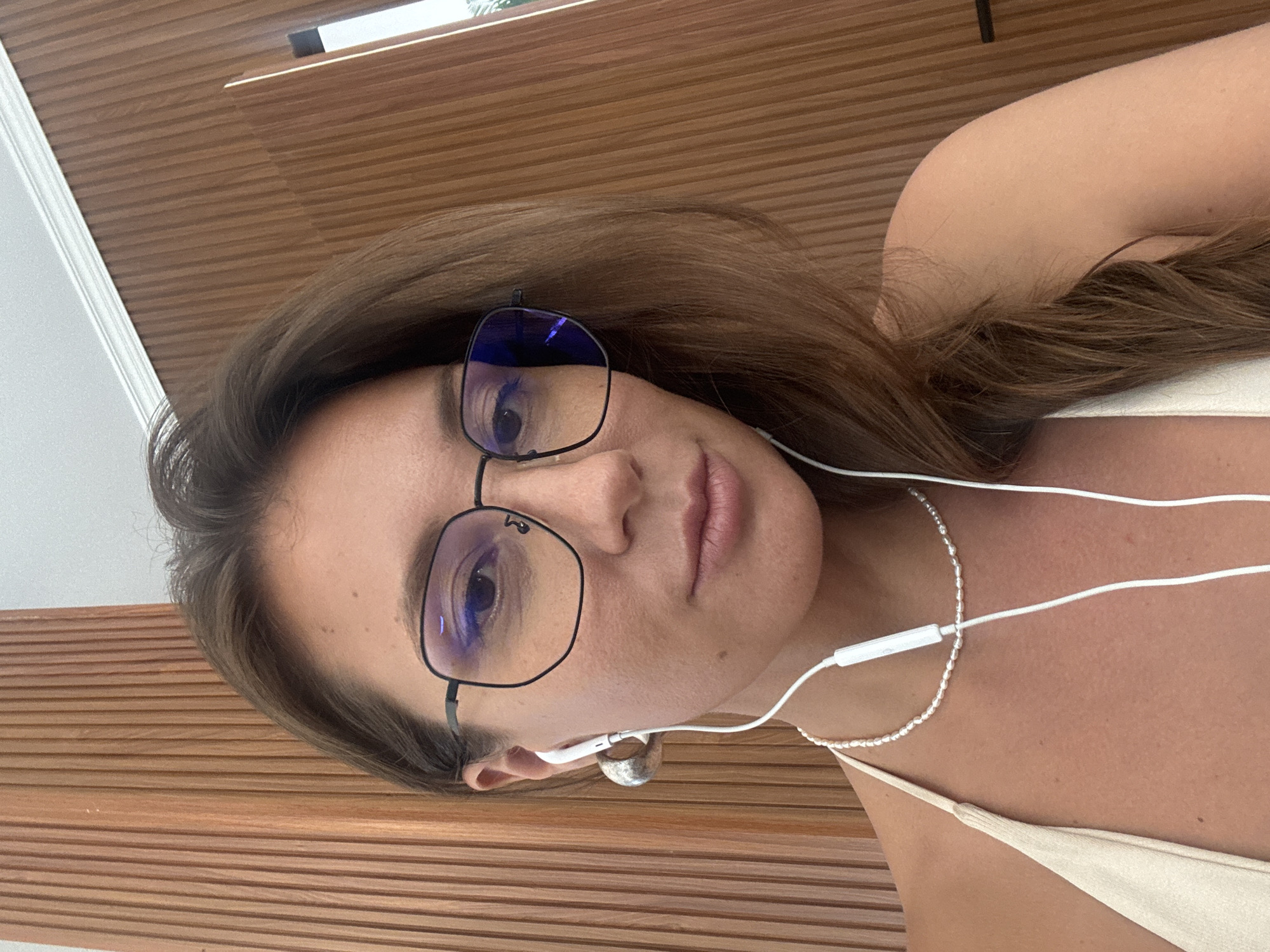 A woman with light brown hair, wearing black-rimmed glasses, is shown in a close-up selfie. She has fair skin, is wearing a silver necklace and a light-colored top, and has white earphones in her ear. The background consists of horizontal wood panels, possibly a wall or ceiling. There is no direct evidence in the image to confirm the location is Denpasar, Indonesia.