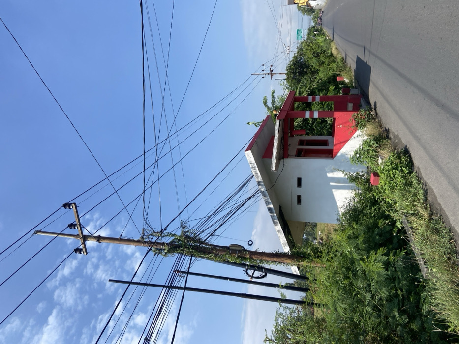 A small, white building with a red-striped roof and porch is situated on the right side of a paved road in Mojosari, Indonesia. The building is located at the edge of the road and surrounded by green vegetation. A power pole with multiple electrical wires extending from it is positioned directly in front of the building. The road extends into the distance, where a few vehicles and people can be seen traveling in the opposite direction. The sky is clear and blue, with only a few wispy clouds visible. The overall scene is one of a quiet, rural setting.