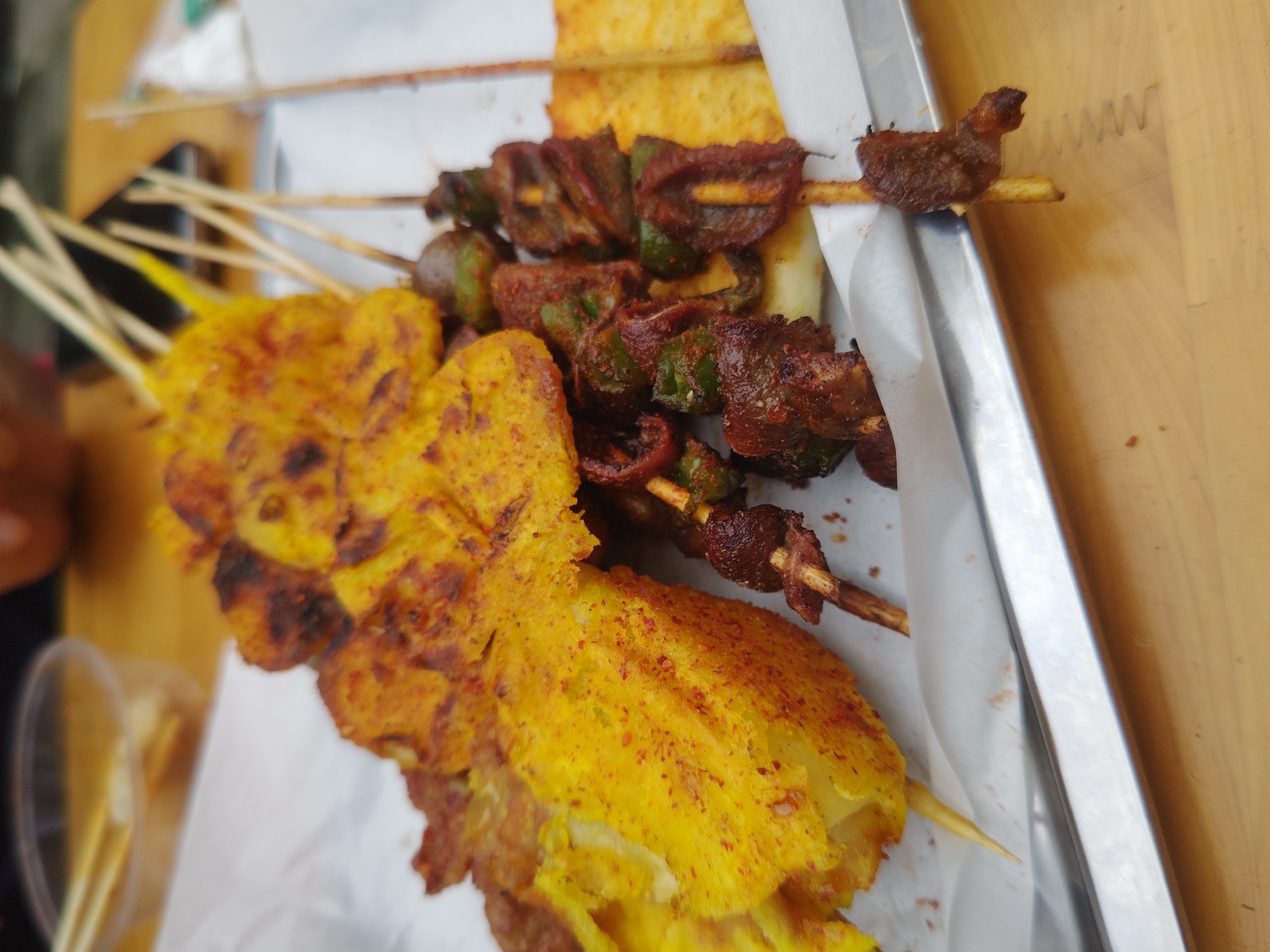 The image shows a tray of skewers, likely food items, in what appears to be an outdoor setting. The skewers are arranged on a white paper liner on a metallic tray. There are two types of skewers: one with yellow-coated food, possibly potato or tofu, and another with darker, reddish-brown meat interspersed with green vegetables, likely peppers. The skewers are all on wooden sticks. The tray appears to be on a wooden surface. The setting appears to be in the city of Chongqing High-tech Zone, China.