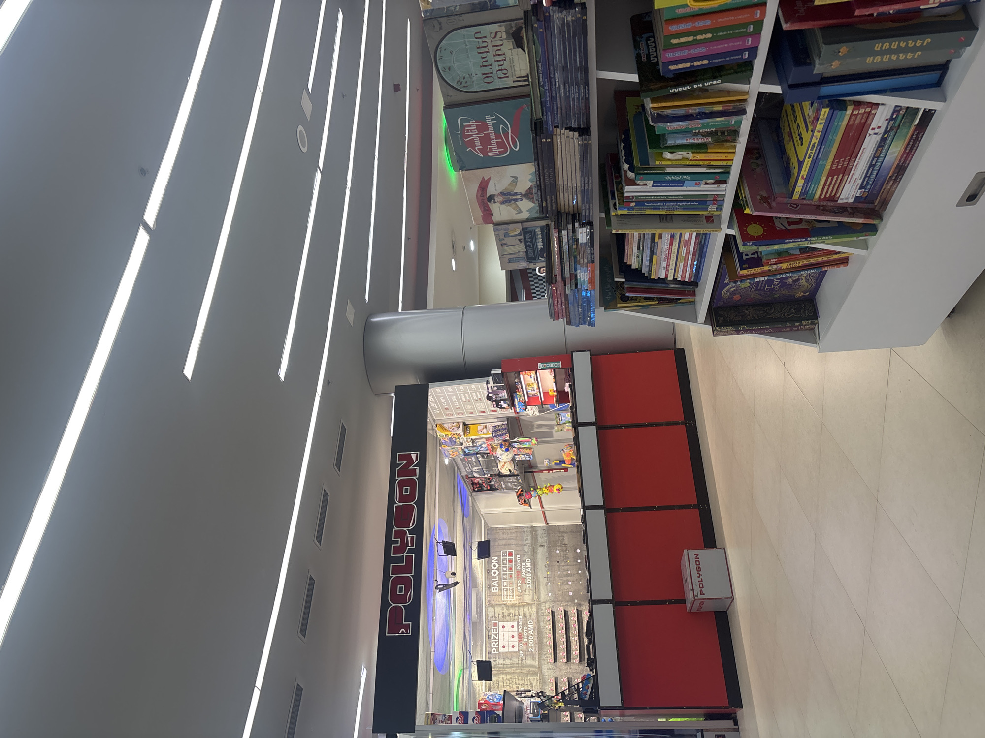 The image displays an indoor commercial setting, likely within a shopping mall or large store in Yerevan, Armenia. The scene is brightly lit by numerous long, linear ceiling lights.
On the left, a prominent black sign with red lettering reads "POLYGON," identifying an entertainment or gaming zone. Below the sign, an arcade-style area features targets and a balloon game. Visible text indicates "PRIZE UP TO 100 POINTS 2,000 AMD" and "BALOON UP TO 6 POINTS 3,000 AMD," referencing the Armenian Dram currency. There appear to be a few indistinct figures within the gaming area, possibly people engaging with the games.
On the right, a large white bookshelf is densely filled with books. Many of the book titles are in Armenian, providing a clear cultural and geographical cue. Recognizable Armenian titles include "ՕԼԻՎԵՐ ԹՎԻՍՏ" (Oliver Twist), "Համլենի Սրնգահարը" (The Pied Piper of Hamelin), "Վիլհելմ Թել" (William Tell), and "ԱՌԱԿՆԵՐ" (Fables/Proverbs). A white box with the "POLYGON" logo is visible on the light-colored tiled floor.
The scene depicts a modern, well-lit environment that combines leisure activities (arcade) with a retail book section. The time of day is not discernible due to the indoor lighting. No specific activities are clearly underway, nor are any emotions conveyed. The Armenian text and currency strongly confirm the location as Yerevan, Armenia.