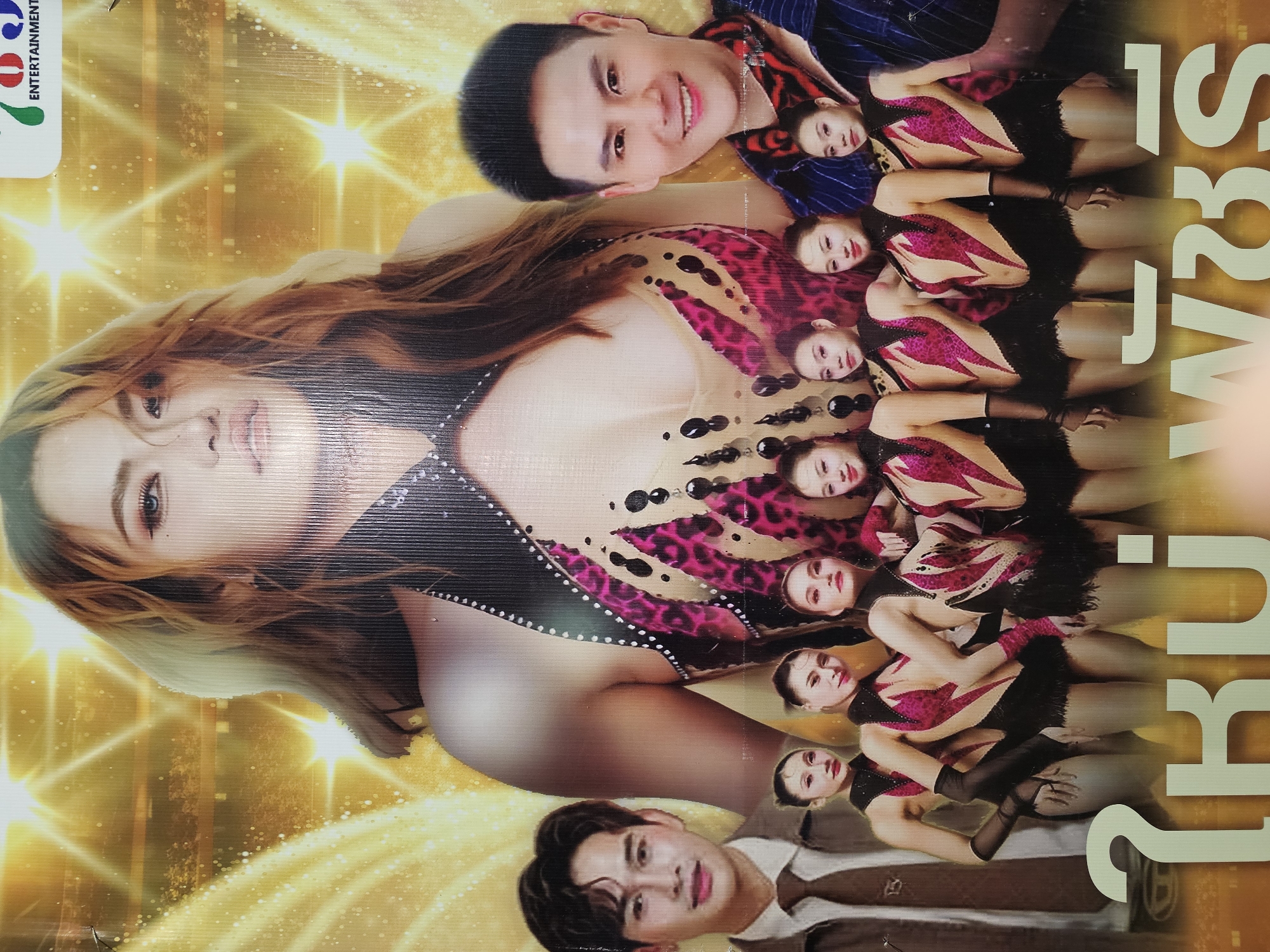 This image is a poster for an event, likely a performance or a show, featuring several individuals. In the foreground, there are several young women dressed in matching, sparkly, black and pink outfits that appear to be dance costumes. They are standing in a row, facing the viewer. Behind them, on the left, is a man in a brown jacket and tie, and on the right is another man in a dark blue shirt and a red and black scarf. Dominating the upper portion of the poster is a close-up of a woman's face and shoulders, with her hair styled and makeup applied for a dramatic look. The background is a bright, golden, star-like pattern, suggestive of a stage or a celebratory atmosphere. At the bottom of the poster, large Thai text is visible. The poster's overall aesthetic is glamorous and attention-grabbing, typical of entertainment promotion. The text at the top right corner of the poster contains the word "ENTERTAINMENT," suggesting the context of the event. Without specific landmarks visible, the location in Bangkok, Thailand, cannot be pinpointed but can be inferred from the Thai text on the poster and the likely context of such promotional material being displayed in a public space within the city.
