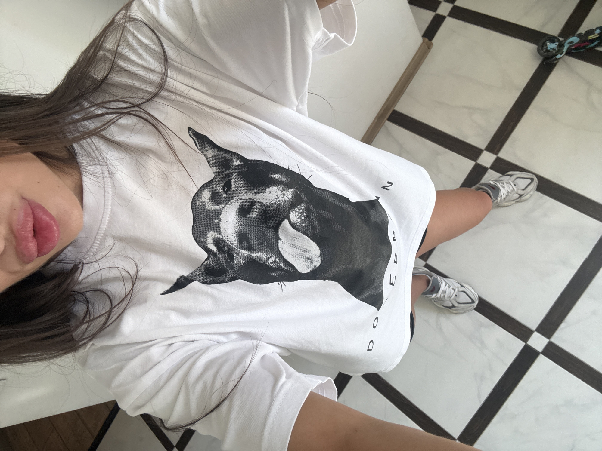 The image shows a woman standing indoors on a tiled floor. She is wearing a white t-shirt with a black and white graphic of a Doberman dog sticking its tongue out, a pair of black shorts, and silver athletic shoes. Her long, dark hair is visible. The floor is made of white tiles with black diamond-shaped insets. The lighting appears to be natural or from overhead lights. The setting is an interior space, possibly a home or a studio, with a wall visible to the right. There is no indication of the specific location within Chișinău, Moldova.