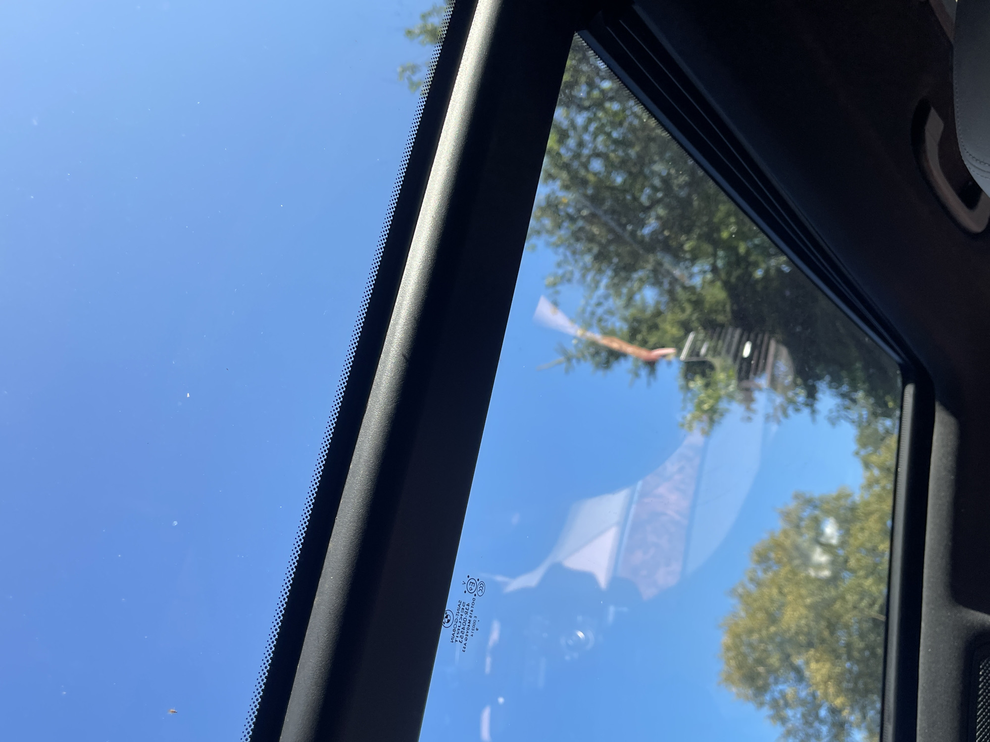 The image is a low-angle, close-up shot looking up through an open sunroof of a car. The sky is a clear, bright blue. Through the sunroof, the tops of green trees are visible, suggesting an outdoor setting. There are some small white dots scattered across the sky, which could be dust or other atmospheric particles. A black frame surrounds the sunroof opening, and the interior of the car, including part of a gray seat and a handle, is visible at the bottom of the frame. On the glass of the sunroof, there is a reflection of something, possibly part of the car's interior or an object outside. There is also some faint text visible on the glass, which appears to be manufacturer information or safety certifications. The scene is likely from a car parked or moving in a location with trees and a clear sky, such as a park or a residential area. There is no identifiable information in the image that places it specifically in Tallinn, Estonia.