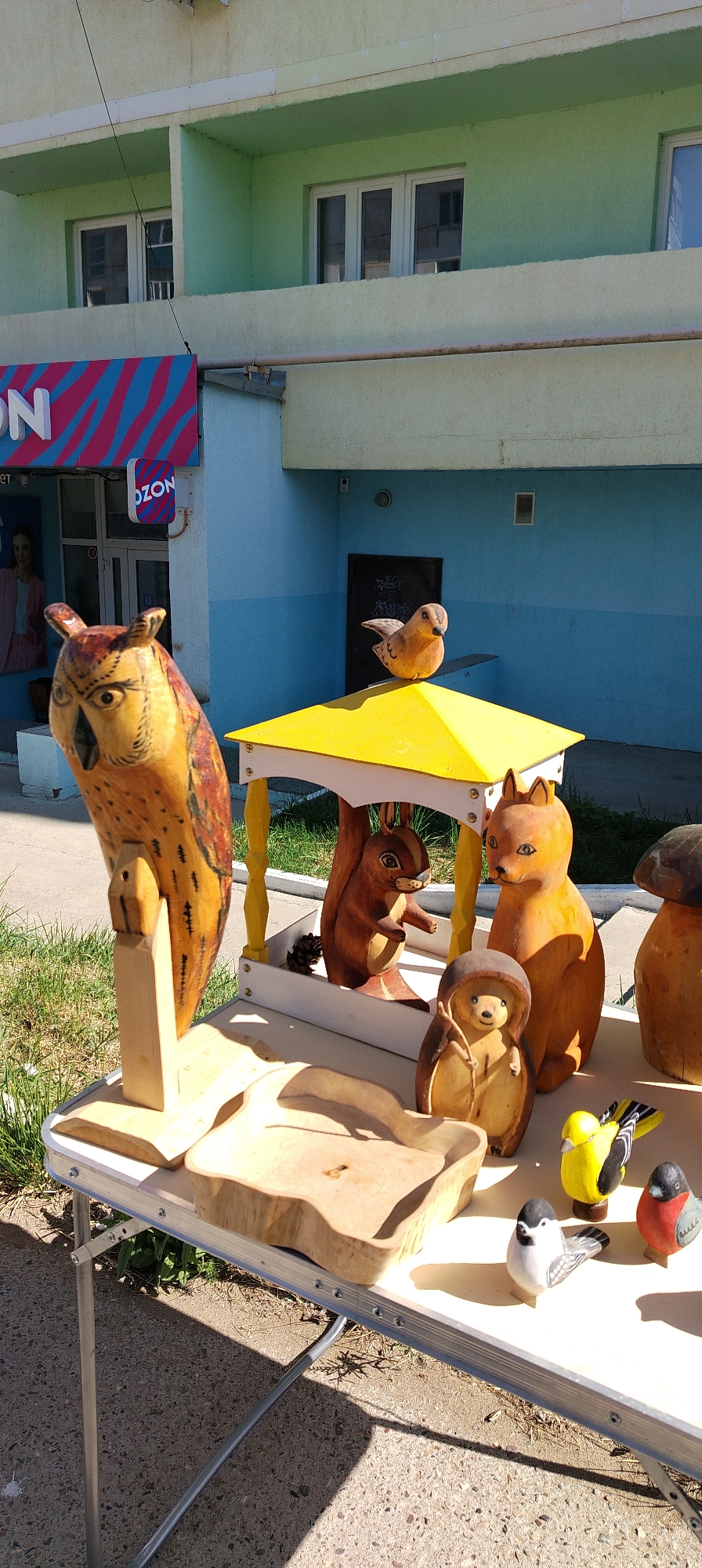 The image depicts an outdoor market stall in the city of Chaikovsky, Russia, featuring an assortment of carved wooden figurines. The stall is set up on a folding table on a concrete surface, with a grassy area and a building in the background. The building appears to be a commercial establishment with a light yellow and blue facade, featuring windows and a green balcony. A sign for "OZON" is visible on the building.
The main subjects of the image are the wooden carvings. Prominently displayed is a large, intricately carved wooden owl with detailed feather patterns and large eyes, standing on a wooden base. To its right, under a bright yellow canopy structure, are carved figurines of woodland creatures. A squirrel is perched within the canopy, while a fox-like animal stands next to it. In front of these, a smaller hedgehog-like figurine is positioned.
On the table, further to the right, are several smaller carved birds. One is a vibrant yellow bird with black and white markings, and another is a red and grey bird. A black and white bird with a gray back and wings is also present. In the foreground, a natural wood carving with an irregular shape, possibly a bowl or decorative piece, is placed on the table. Another carved wooden mushroom is visible on the far right.
The lighting suggests it is daytime, with strong shadows cast by the objects on the table. The overall scene captures a display of handcrafted wooden art being sold outdoors.