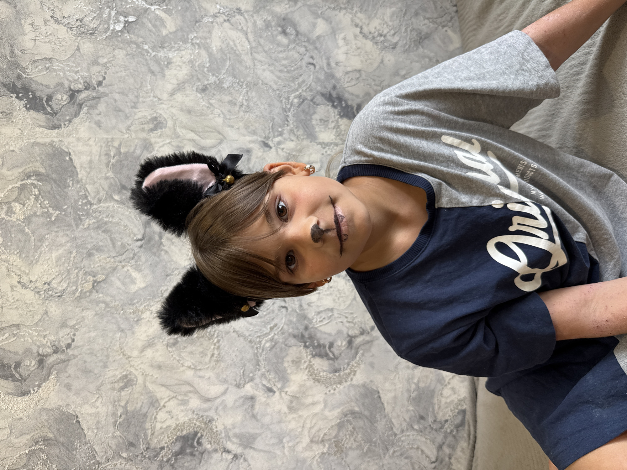 In this media file, a young person is depicted lying on a textured gray surface that appears to be a bed or sofa. The person is wearing a costume, complete with black furry cat ears with pink inner lining, adorned with a black ribbon and a small bell. They also have face paint that resembles a cat's whiskers and nose. The person is wearing a dark blue t-shirt layered with a gray long-sleeved shirt over it. The gray shirt has white text on the sleeve that appears to be a logo or brand name, with "NTS" and "NTS" clearly visible, and part of what looks like "A R" and "RY" also present. The overall mood seems playful and whimsical, possibly for dress-up or a costume event. The lighting suggests an indoor setting, possibly during the daytime. The background is a textured, gray, patterned surface. The location is identified as Rusanovo, Russia.