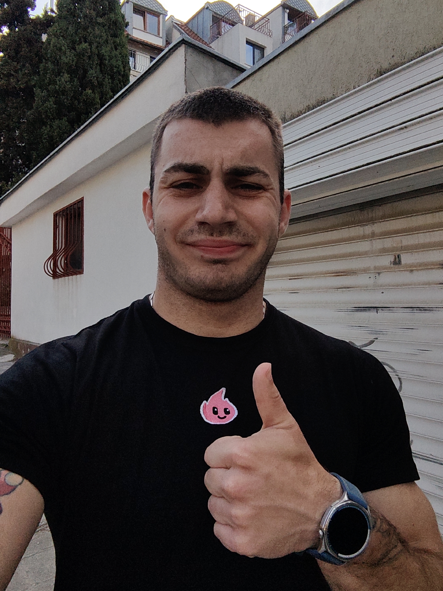 A man is standing outdoors in Varna, Bulgaria, giving a thumbs-up gesture. He is wearing a black T-shirt with a small, cartoon-like pink flame with a smiling face embroidered on it. He has short, dark hair and a light beard. On his left wrist, he is wearing a smartwatch with a blue band. His left arm shows a tattoo.
Behind him is a white building with a red, decorative window grate. To the right, there's a commercial building with a closed, horizontal-slatted metal security door. Further back, the scene reveals other buildings with dormers and wrought-iron balconies. A tall, dark green tree is visible on the left side of the frame. The image appears to be taken during daylight hours.
