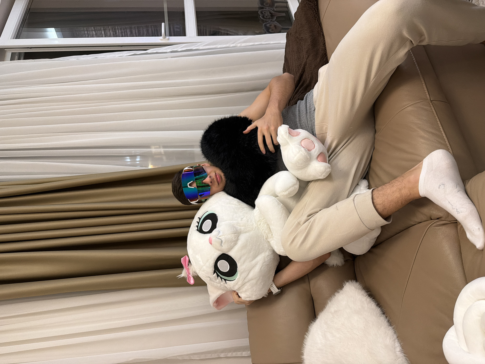 A person lies on a brown leather couch wearing beige pants, a grey shirt, and multicolored reflective sunglasses. They are surrounded by various plush items, including a large white cat with a pink bow, a black furry pillow, and a white paw-shaped plush. The person appears to be holding the plush cat. The location appears to be indoors, possibly a living room, with white curtains and a window in the background. The setting is Unknown, Unknown.