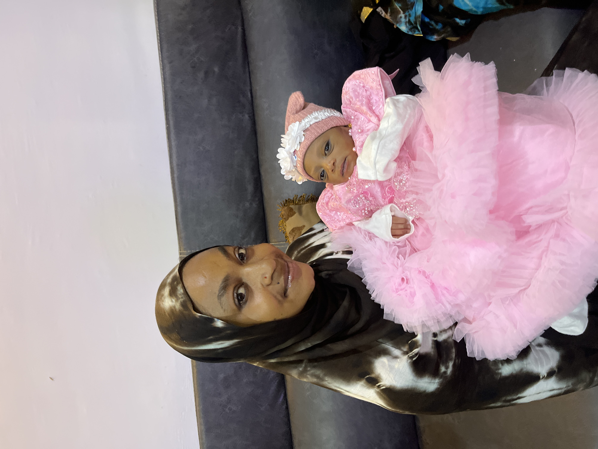 A woman wearing a dark and tan patterned hijab is holding a baby dressed in a pink ruffled dress and hat. The woman is smiling at the camera, and the baby is looking towards the camera. They are seated on a dark leather couch. The background is a plain, light-colored wall. The overall scene appears to be indoors. There are no visible elements that indicate the location is specifically within the city of Jos, Nigeria.