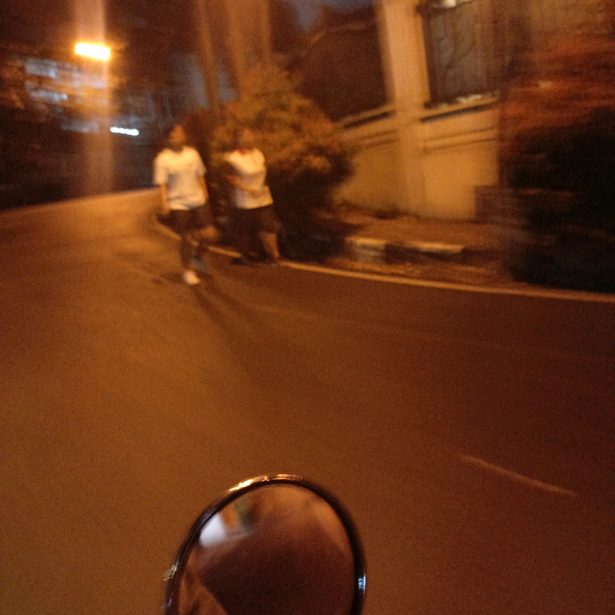 At night, on a city street in Salatiga, Indonesia, a person on a motorcycle is recording a video. The camera is focused on the road ahead, which is illuminated by streetlights and the headlights of passing vehicles. In the background, two people can be seen walking on the sidewalk. One of them is wearing a white t-shirt and the other is wearing a dark t-shirt. The street is paved with asphalt and there are trees and buildings on either side. The overall scene is blurry due to the low light conditions and the movement of the motorcycle.