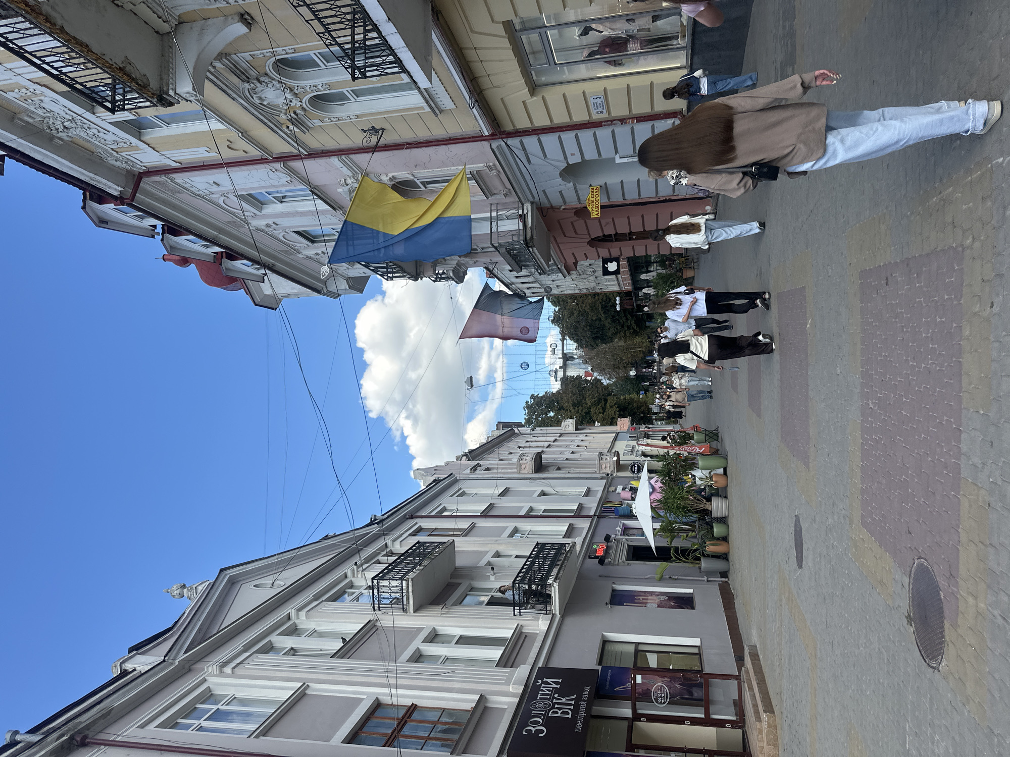 The image captures a sunny day on a pedestrian street in Ternopil, Ukraine, likely in the historic city center. The street is paved with patterned cobblestones and lined with multi-story buildings showcasing traditional European architecture with ornate facades. The buildings have large windows, balconies with decorative iron railings, and a mix of light-colored paint schemes. Ukrainian flags are prominently displayed on some of the buildings, a common sight in the country.
Several people are walking along the street, dressed in casual attire. The individuals in the foreground are walking away from the camera, while others are seen in the distance. Some storefronts are visible along the street, including one with a sign featuring Ukrainian text. There are also potted plants and outdoor seating areas in front of some establishments, suggesting the presence of cafes or restaurants. The sky is blue with scattered white clouds, and the overall atmosphere appears lively and bustling.
