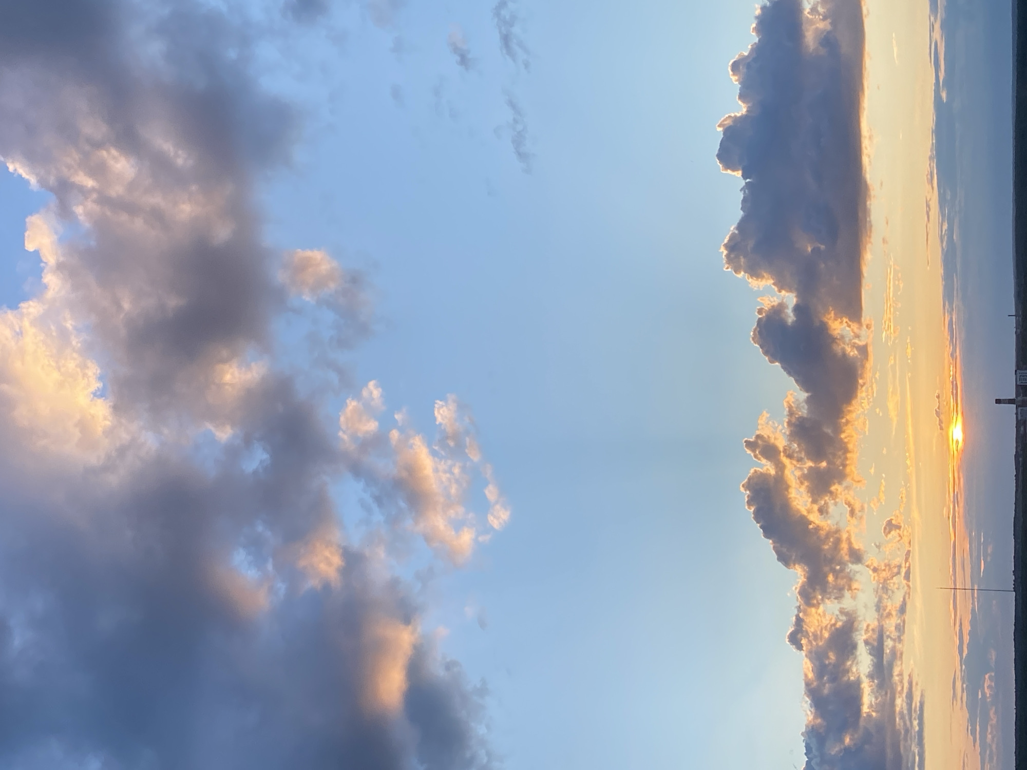The image captures a beautiful and expansive sunset sky over a distant horizon. The sun, a bright golden orb, is visible low on the right side of the frame, partially obscured by clouds and casting a warm, luminous glow across the scene.
The sky features a striking contrast between clear blue areas and dramatic cloud formations. Towards the left, a large mass of darker, greyish-blue clouds with soft, pinkish-gold edges occupies a significant portion of the sky, indicative of the warm light hitting their undersides. In the center, patches of clear, light blue sky are visible. On the right, closer to the setting sun, another extensive cloud formation is brightly illuminated from behind and below, with its edges and undersides glowing intensely in vibrant oranges, yellows, and gold, standing out against the dimmer sky and darker cloud bodies.
The horizon line stretches across the lower part of the image, appearing as a dark, flat band. Along this distant line, faint, dark silhouettes of what might be land features, trees, or possibly structures can be vaguely discerned, particularly on the far right where a thin vertical line, perhaps an antenna or pole, stands against the bright sunset.
The time of day is clearly sunset, characterized by the low sun, the warm, golden and orange hues dominating the sky, and the soft, indirect lighting on the clouds. The scene conveys a sense of vastness, tranquility, and natural beauty. There are no people, animals, or prominent man-made objects in the foreground or midground. No visible text is present. The general setting is an open sky over a landscape, consistent with the location context of Izhevsk, Russia.