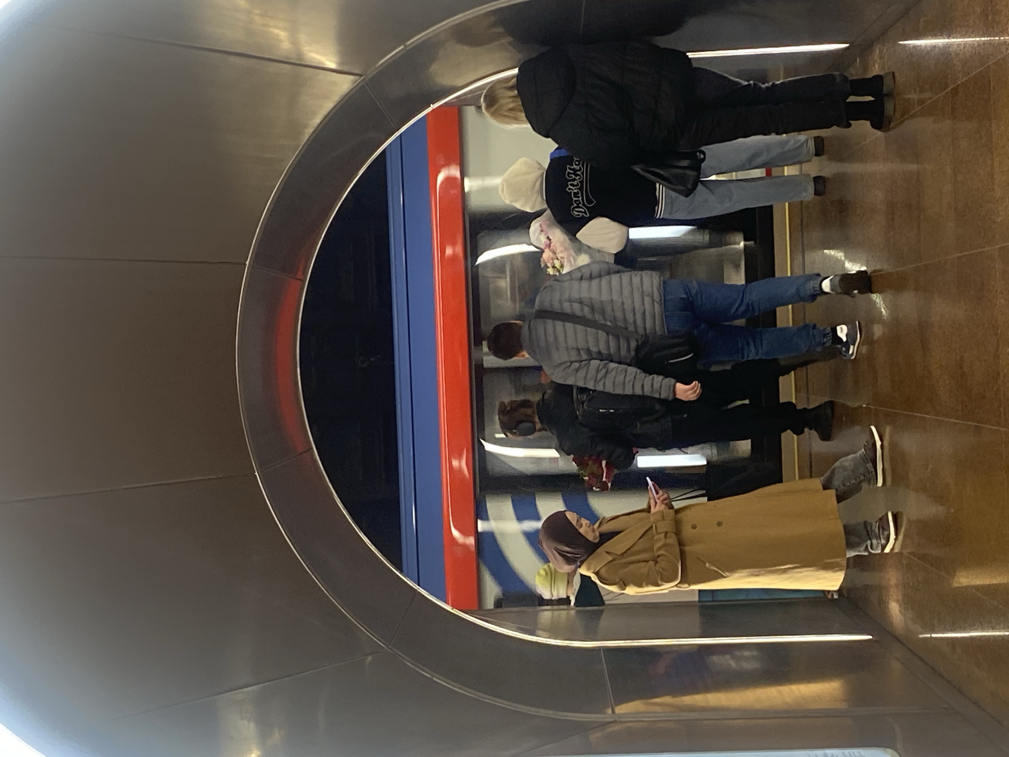 The image shows a group of people standing in a subway station. A train is visible on the right side of the frame, with blue and red stripes. The people are standing on a brown tiled floor, and the station has a metallic, curved wall on the left. One woman in the foreground wears a brown coat and a headscarf and is looking at her phone. Other people in the background are wearing casual clothing such as jackets, jeans, and sneakers. Some appear to be waiting for the train. The location appears to be in an underground subway or metro station.