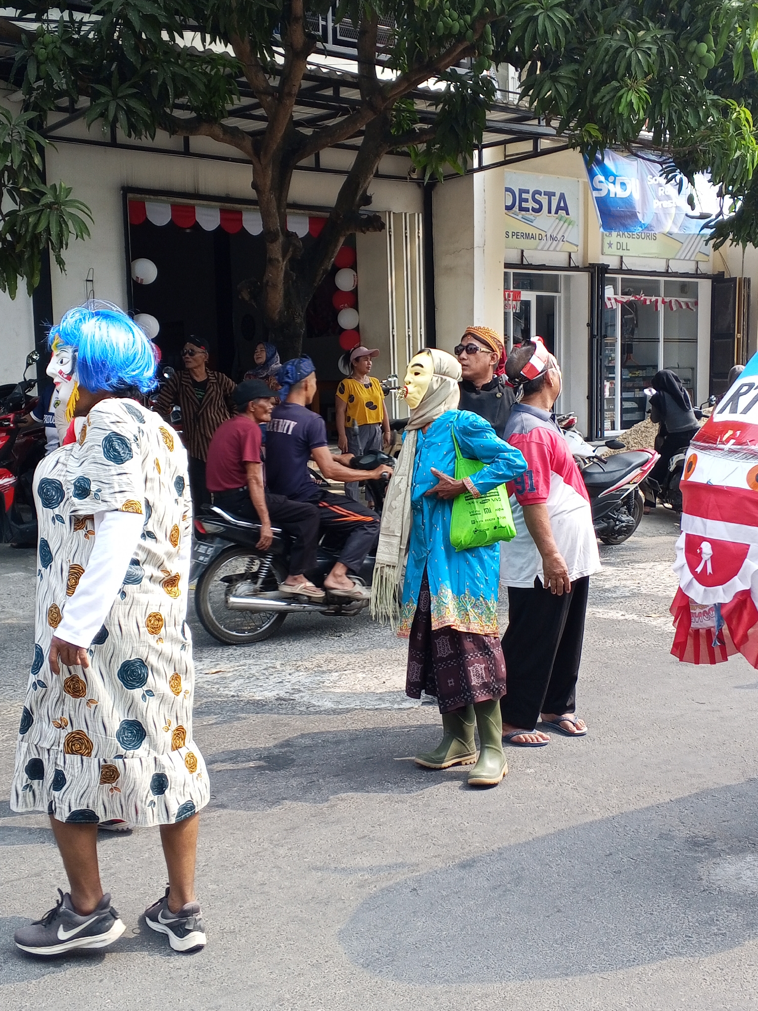 A group of people are gathered on a street in Ciruas, Indonesia, in front of a building with shops. In the foreground, a person wearing a blue wig, a white dress with a floral pattern, and grey sneakers stands facing left. To their right, a person wearing a blue tunic, a light brown headscarf, a tan mask, and green boots is carrying a green bag. Another person in a red and white shirt and black pants stands next to them. In the background, a person in a maroon shirt rides on a motorcycle, with another person wearing a navy blue shirt behind them. Several other individuals are visible standing or sitting around the street, some of whom are wearing masks. The environment appears to be a commercial street with trees and buildings lining the road. Red and white decorations are visible on the buildings.