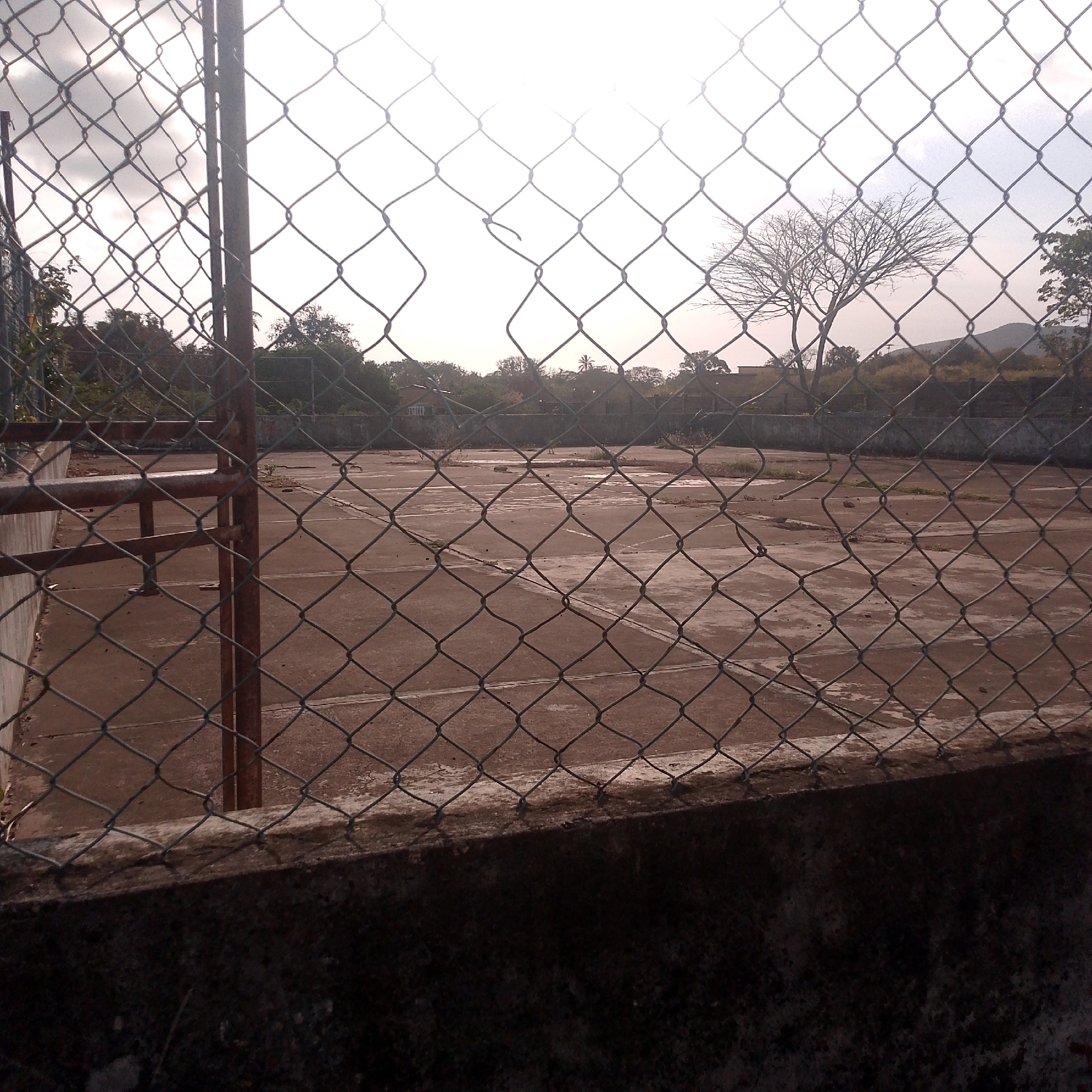 The image shows an abandoned tennis court in Altagracia, Venezuela. A chain-link fence surrounds the court, with the view partially obstructed by the fence itself. The court surface is cracked and weathered, with scattered debris. In the background, there are trees and a distant mountain. No people are visible.