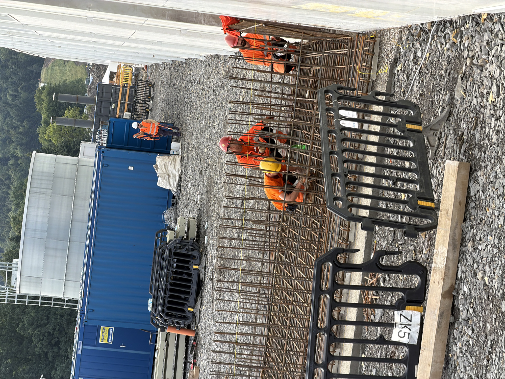 The image shows a construction site in Janowice, Poland. In the foreground, several construction workers are actively engaged in reinforcing a concrete structure with steel rebar. Three workers are visible, all wearing orange high-visibility vests and hard hats. One worker in the center, wearing a yellow hard hat, is crouched down and appears to be securing rebar. To his left, another worker in a red hard hat is also working with the rebar. To the right, a third worker in a red hard hat is facing away from the camera, also working on the reinforcement. In the mid-ground, there are blue shipping containers, some stacked. A man in an orange vest and a white hard hat is standing near one of the containers, holding a harness. In the background, a large, cylindrical structure, possibly a water tank or silo, is visible, along with trees and a wooded hillside. The ground is covered with gravel and construction debris. Black plastic barriers are placed to cordon off areas of the site. A wooden plank lies on the ground in the lower left corner, with a white tag bearing the inscription "ZK5" attached to one of the barriers. The scene appears to be an active construction area with ongoing work on a foundation or structural element.