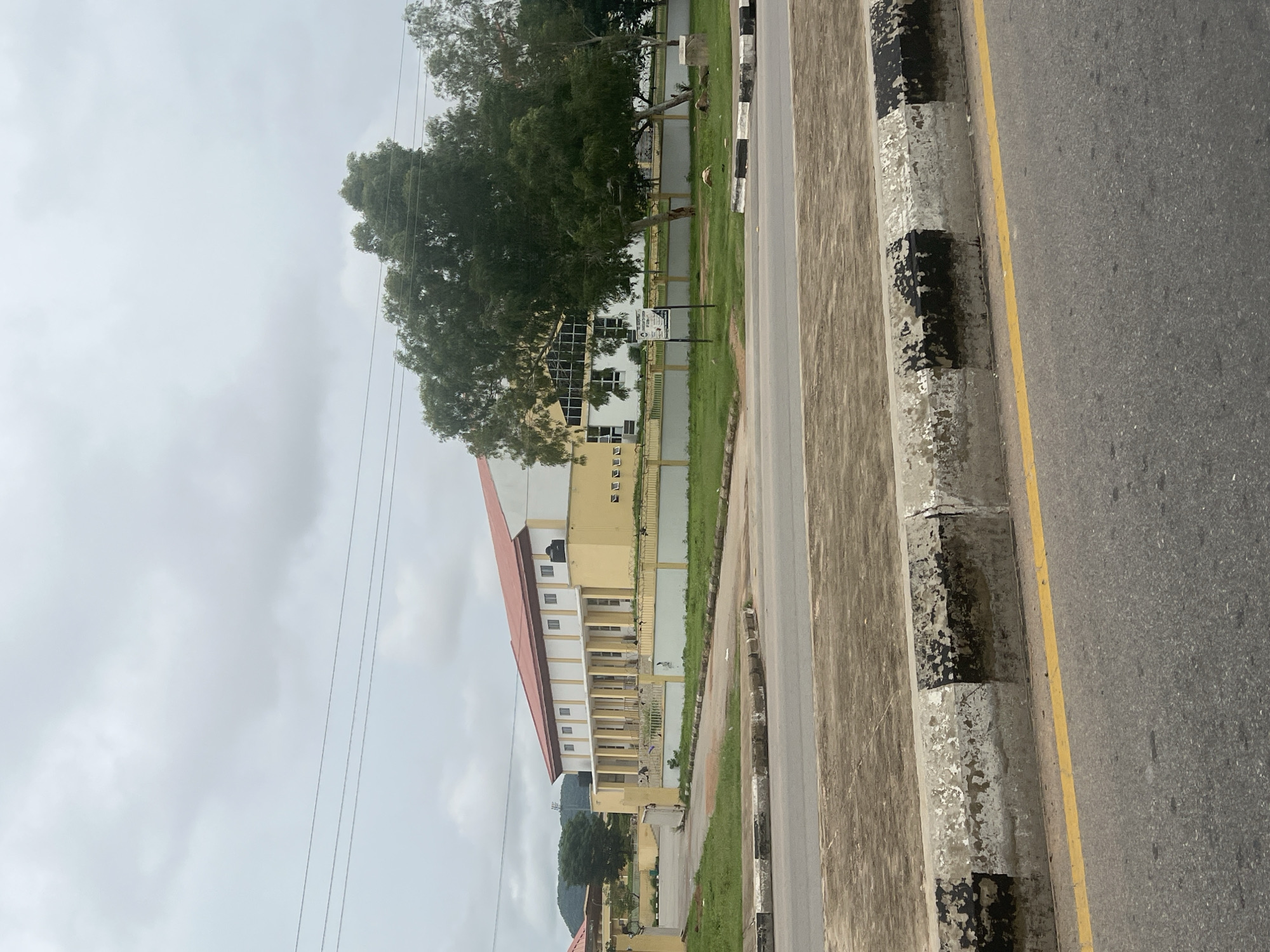 The image depicts a large, light yellow and white building with a red roof, likely a public institution or educational facility, located in Howayi, Nigeria. The building is situated behind a low yellow and white fence with a metal railing. Several trees, including a prominent mature tree with dense foliage, are in front of and to the side of the building. A road runs in the foreground, with a concrete curb marked with black and white stripes, showing signs of wear. Overhead power lines are visible against a cloudy sky. The scene appears to be on an overcast day, with natural light. A signpost is visible on the grass in front of the building, but its text is illegible. The overall environment suggests an urban or semi-urban setting.