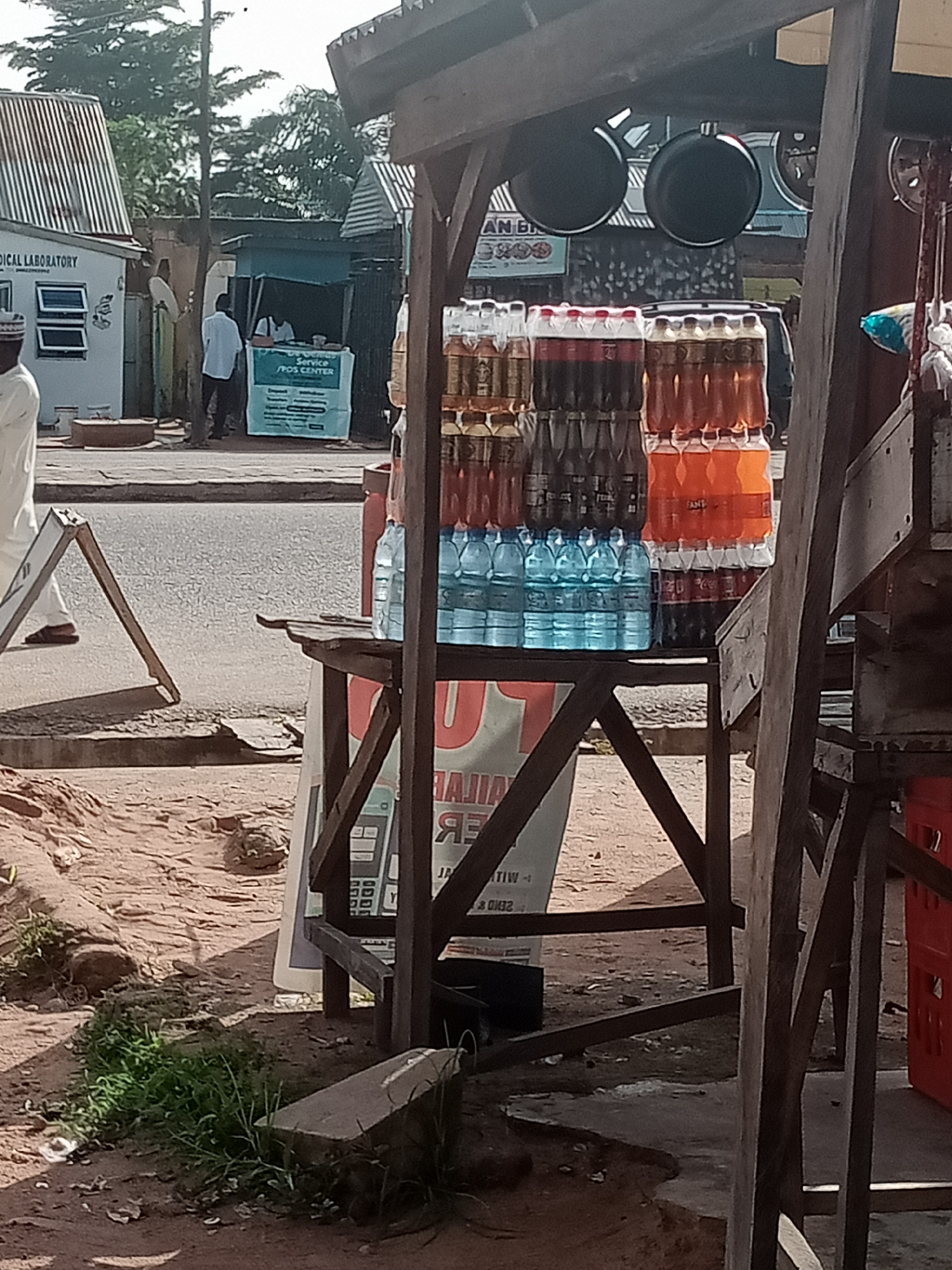 The image depicts an outdoor scene on a street in Jalingo, Nigeria, likely near a market or commercial area. In the foreground, a makeshift wooden stand displays an assortment of bottled beverages. Stacked in rows, there are several bottles of clear liquid, dark-colored soda, and orange-colored drinks. The stand is constructed from rough wooden planks, with some supporting beams visible.
To the left of the stand, a busy street or road is visible, with a sidewalk and some sand and dirt on the ground. A white A-frame sign stands on the sidewalk. Further in the background, a building with a corrugated metal roof and the words "MEDICAL LABORATORY" is seen. Adjacent to it, a smaller stall with a blue awning appears to be a service point or POS center, with people standing and interacting. The overall environment suggests a typical street scene in a Nigerian city, characterized by commercial activity and a blend of basic infrastructure. The lighting indicates it is daytime, with shadows cast by the structures and objects.