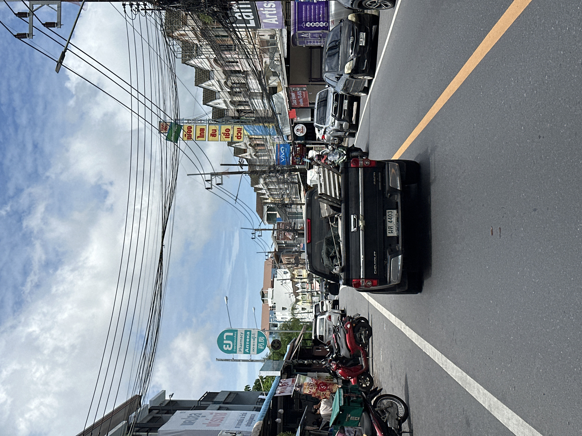 A medium shot captures a street scene in an urban environment under a cloudy sky. The perspective is from a vehicle traveling on the right side of a paved road. The road is divided by a white lane marker on the left and a yellow dashed line on the right.
In the foreground, the rear of a black Chevrolet pickup truck is visible, with a license plate reading "ผฉ 4403". The truck bed is laden with what appears to be metal scaffolding. To the left of the truck, several parked motorcycles are visible, including a red scooter.
The street is lined with buildings on both sides. On the left, a row of one-story establishments with awnings creates a market-like atmosphere. Signs in Thai are present, including one for "Pharmacy". On the right, multi-story buildings with pitched roofs line the street, featuring various shop fronts and advertisements. One sign on the right reads "Lam Artistic".
Several vehicles are on the road, including a black sedan with license plate "กร 9812", a silver SUV, and a white sedan further ahead. A motorcycle with a rider is passing the silver SUV. Overhead, a complex network of electrical wires is strung between poles, crossing the sky. The overall impression is a busy street in a Southeast Asian city.