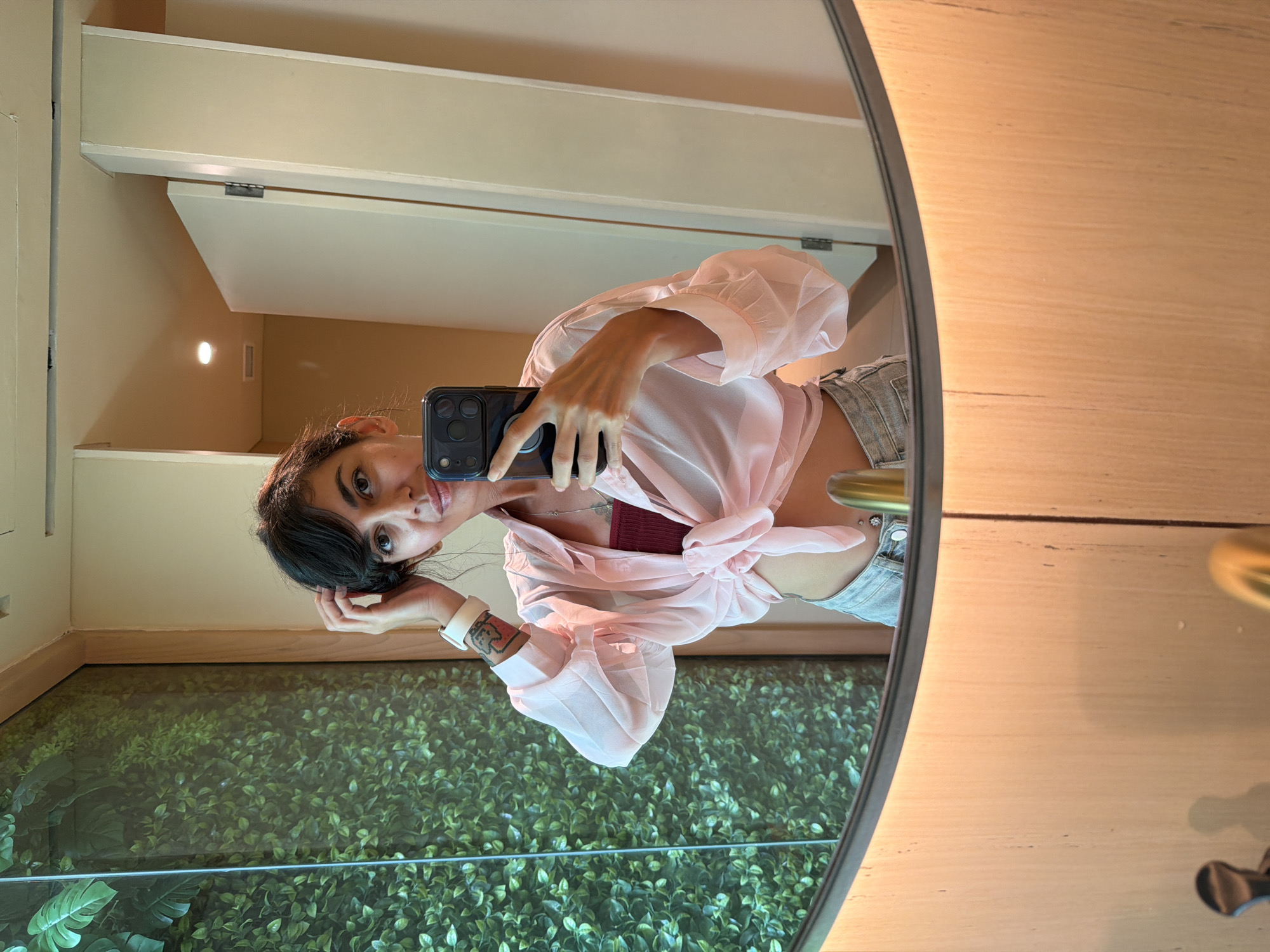 A young woman with dark hair and fair skin is taking a selfie in a bathroom mirror. She wears a pale pink, long-sleeved, tie-front shirt, gray denim shorts, and a burgundy top underneath. Her phone is black, with multiple camera lenses visible. A white smartwatch is on her left wrist, revealing a tattoo. The bathroom setting includes a beige wall, a white door, and a decorative wall of greenery, possibly artificial plants, seen in the mirror. The mirror itself is oval-shaped with a tan wooden frame. Based on the location in North Kuta, Indonesia, the environment suggests a modern, possibly high-end, bathroom in a residence or hotel.