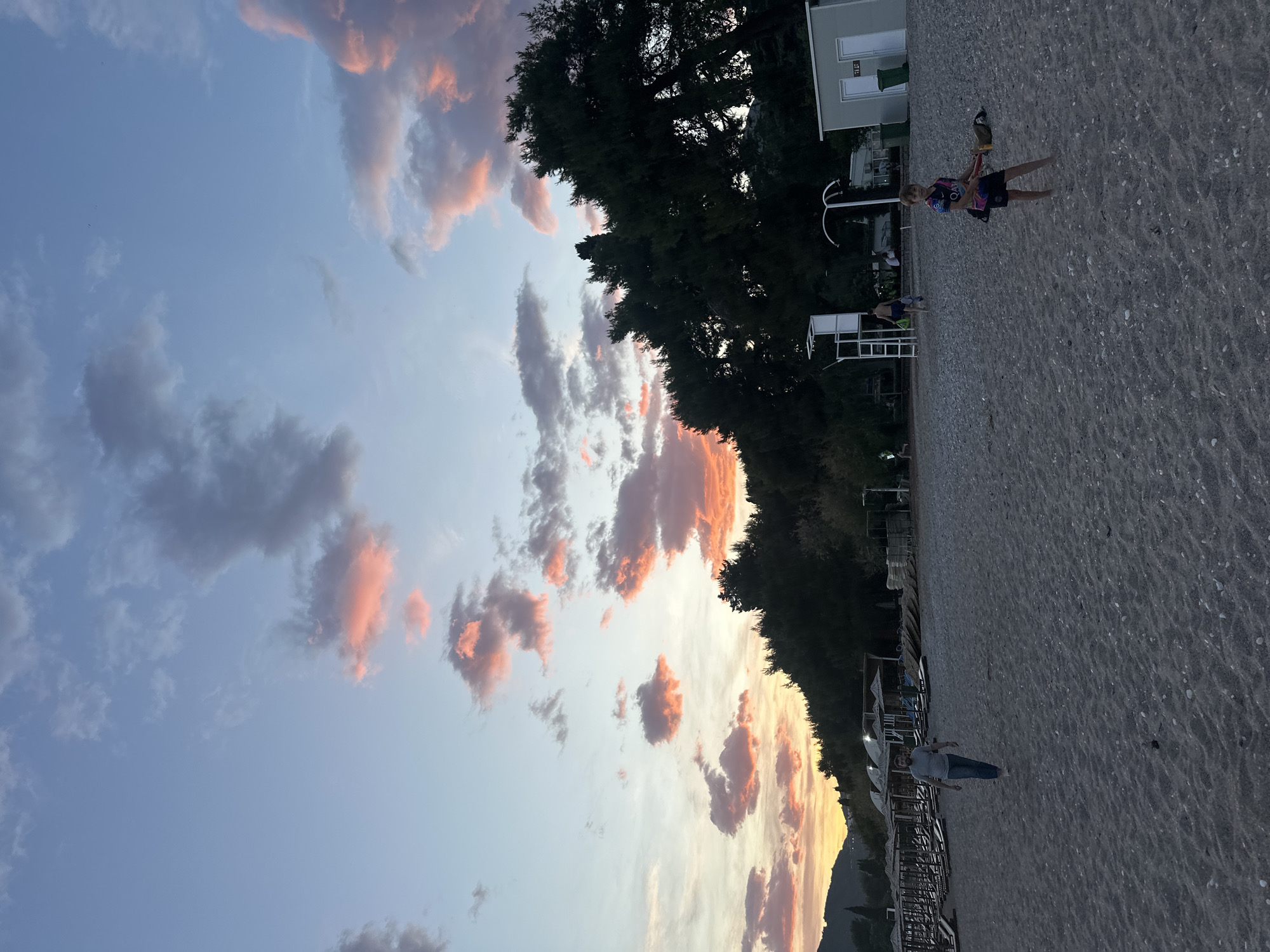 The image depicts a beach scene in what appears to be the city of Budva, Montenegro, during sunset. The sky is filled with numerous clouds tinged with orange and pink hues, suggesting the presence of sunlight. A line of trees borders the beach. Several individuals are visible on the beach. A boy is walking barefoot, carrying objects, and two other individuals appear to be strolling. The beach itself consists of grey sand. Several beach umbrellas are lined on the shoreline. A building or structure is visible near the tree line. The overall scene suggests a tranquil evening on the beach.