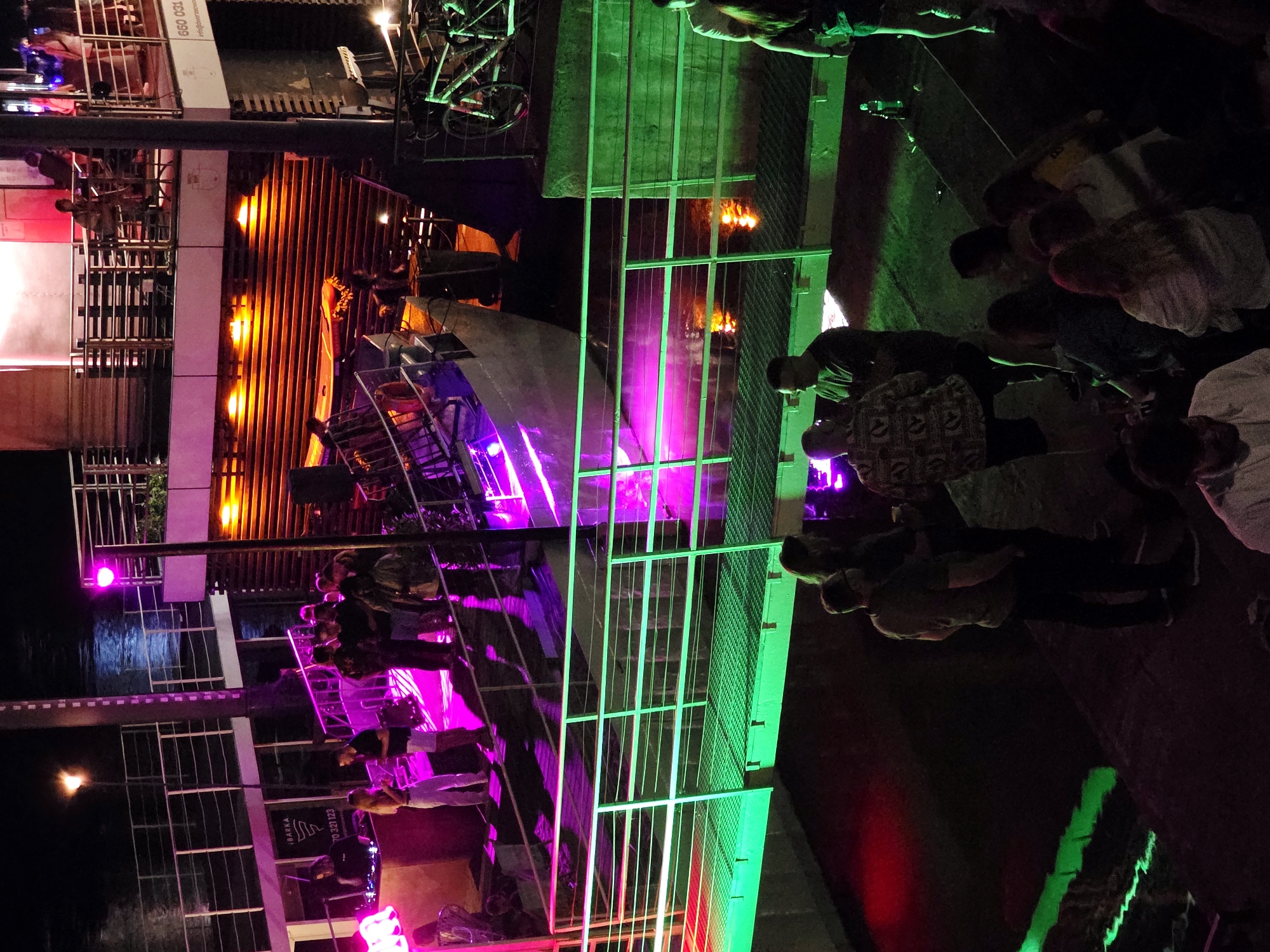 This is a nighttime photograph of a waterfront venue in Warsaw, Poland, likely a club or bar situated on a boat or pier. The scene is illuminated by vibrant pink and green lights, creating a festive and energetic atmosphere.
In the foreground, a group of people are gathered on a walkway. One man, wearing a patterned shirt and dark shorts, is facing away from the camera, while another man in a dark t-shirt and glasses stands beside him. Further back, more individuals are visible, some socializing and others appearing to be part of a crowd.
Above the walkway, a multi-level structure is visible with railings and seating areas. On one of the upper levels, several people are standing and interacting. A DJ booth with equipment is also present, suggesting a musical performance or event is taking place. A bicycle is parked near the structure.
The background features water, reflecting the colorful lights from the venue. The architecture suggests a modern design with industrial elements, typical of contemporary waterfront developments. The overall impression is one of a lively night out in an urban setting.
