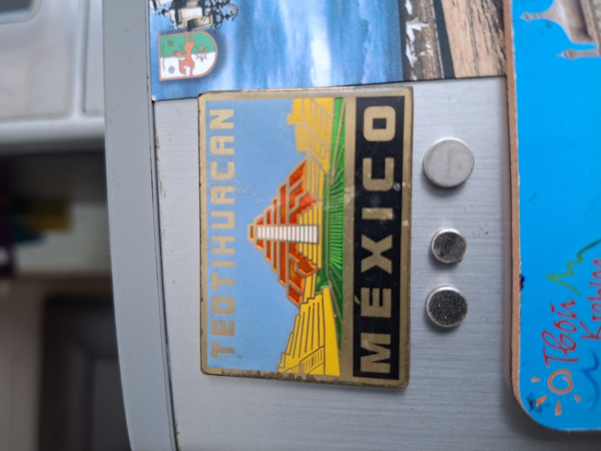 The image shows a close-up of a refrigerator door with several magnets attached. The main subject is a rectangular magnet featuring an image of the Teotihuacan pyramids in Mexico. The magnet has a blue sky background, and the pyramids are depicted in vibrant yellow, red, and green colors. The words "TEOTIHUACAN" are written vertically in blue on the left side of the magnet, and "MÉXICO" is written vertically in black with a gold border on the right side.
To the left of the main magnet, a smaller, oval-shaped magnet is visible. This magnet has a green and white background with a depiction of a red deer-like animal and some foliage. It appears to be a coat of arms or a regional emblem.
To the right of the Teotihuacan magnet, there are three round, silver-colored magnets. Further to the right, a bright blue rectangular magnet is partially visible, adorned with red and orange handwritten text and a drawing of a sunburst.
The overall setting appears to be the interior of a home, likely a kitchen, given the refrigerator. The lighting is even, suggesting indoor illumination. There are no people or active events depicted; the image focuses on the collection of magnets. The magnets suggest a travel theme, with at least two magnets representing destinations in Mexico and another possibly representing a European location. The condition of the magnets indicates they have been attached to the refrigerator for some time, with some minor wear and tear visible on the edges.