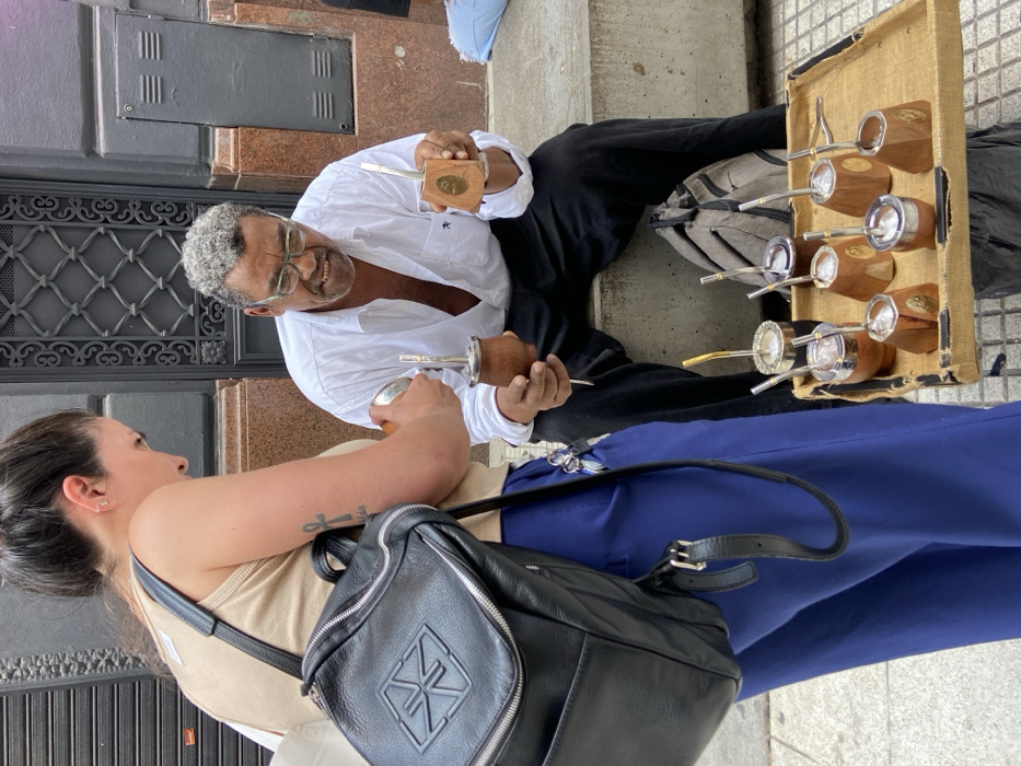 A street vendor sits on the steps, holding two gourds with silver straws, smiling at a woman wearing blue pants and a black purse. To the vendor's right is a display tray with seven similar gourds. The vendor is wearing a white shirt and black pants. The woman has a tattoo on her arm. They are on a city street next to a dark building.