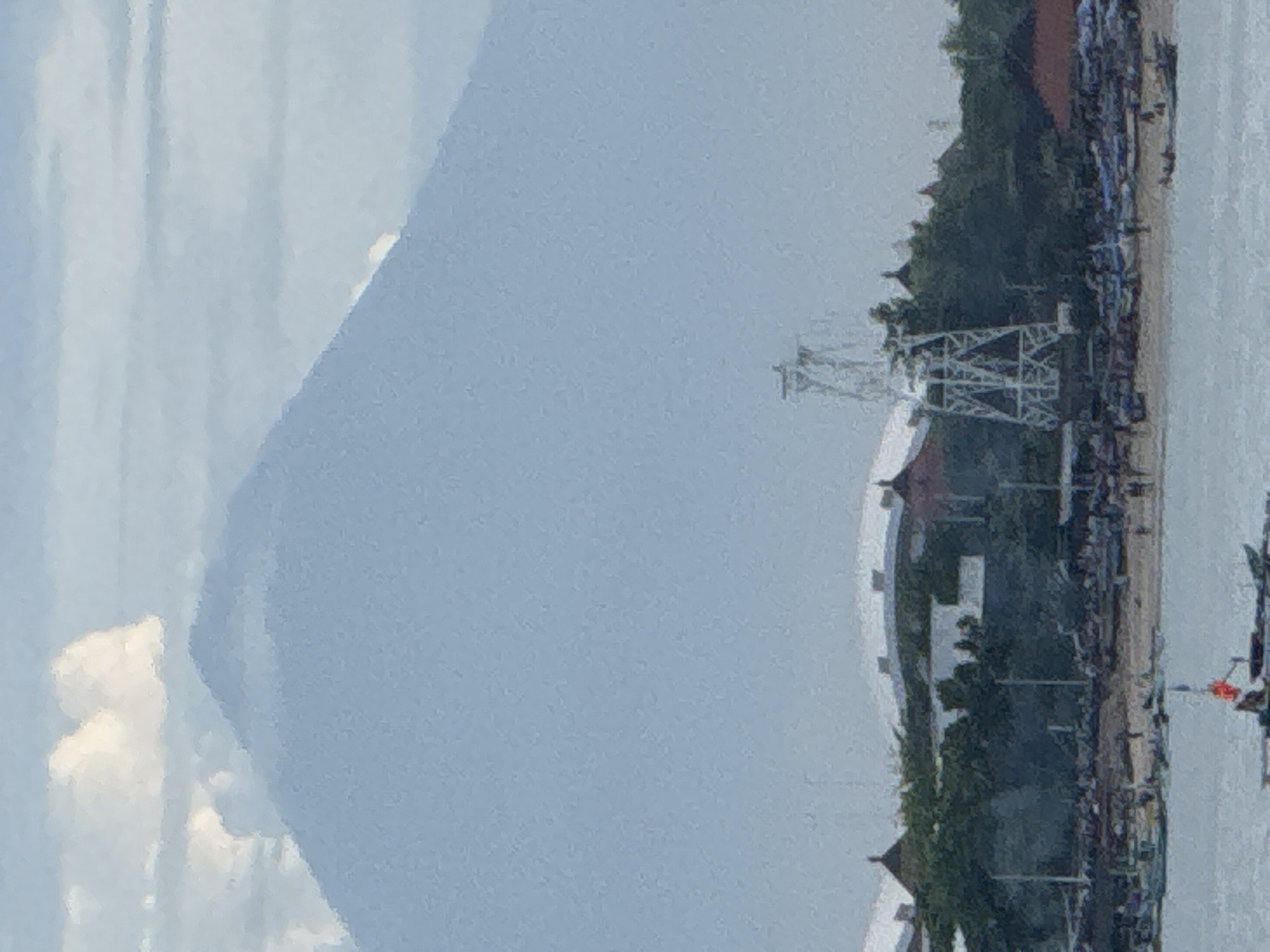 The image shows Mount Agung in Bali, Indonesia, likely taken from the city of Kuta Selatan. The volcano's peak is partially obscured by clouds. In the foreground, there is a coastline with a pier-like structure. Along the beach, there appears to be a dense gathering of boats and buildings. The overall environment suggests a coastal area with a prominent volcanic backdrop.