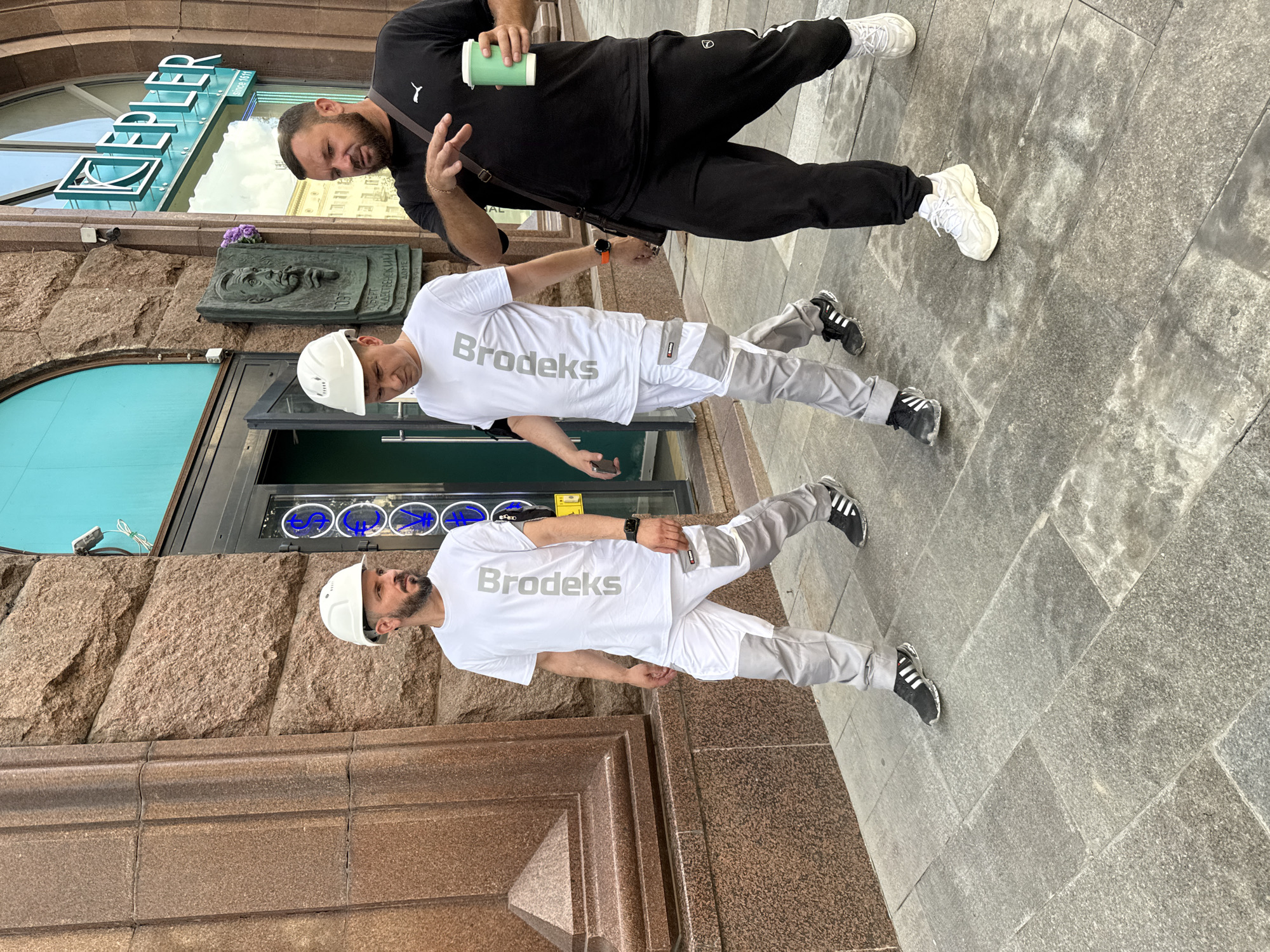 Three men are walking on a paved sidewalk in front of a building with a granite facade. The man on the right is wearing a black t-shirt and black sweatpants, carrying a green cup and a crossbody bag. The two men to his left are wearing white hard hats, white t-shirts with "Brodeks" printed vertically in gray, and white pants with gray accents. They are walking together. The building has a dark entrance with blue circular symbols visible through the glass. A sign with the word "KEPLER" in green is mounted above the entrance. The scene appears to be in Moscow, Russia.