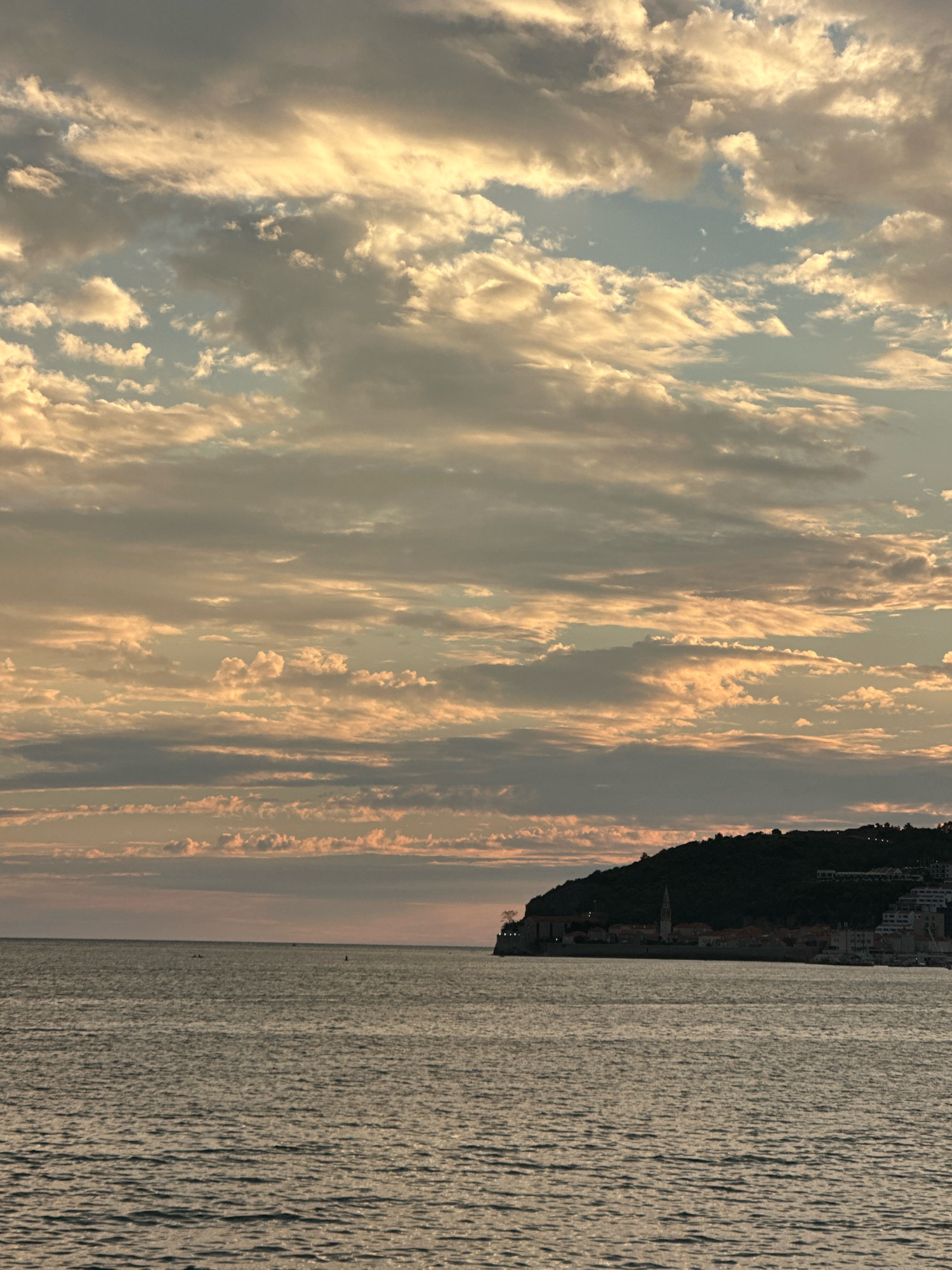 The image shows a body of water, likely the Adriatic Sea, in front of the city of Budva, Montenegro. The sky is filled with a dramatic display of clouds, illuminated by the warm hues of a setting sun. The clouds are layered and varied, with streaks of gold and orange intermingling with shades of gray and blue. In the distance, a dark, tree-covered hill rises from the sea, topped with the buildings of Budva. The city's silhouette is defined by the roofs and structures, including what appears to be a church steeple. The water in the foreground is calm, with gentle ripples reflecting the colors of the sky. A few small boats are visible on the horizon, adding a sense of scale to the vastness of the seascape. The overall impression is one of natural beauty and a serene coastal landscape.