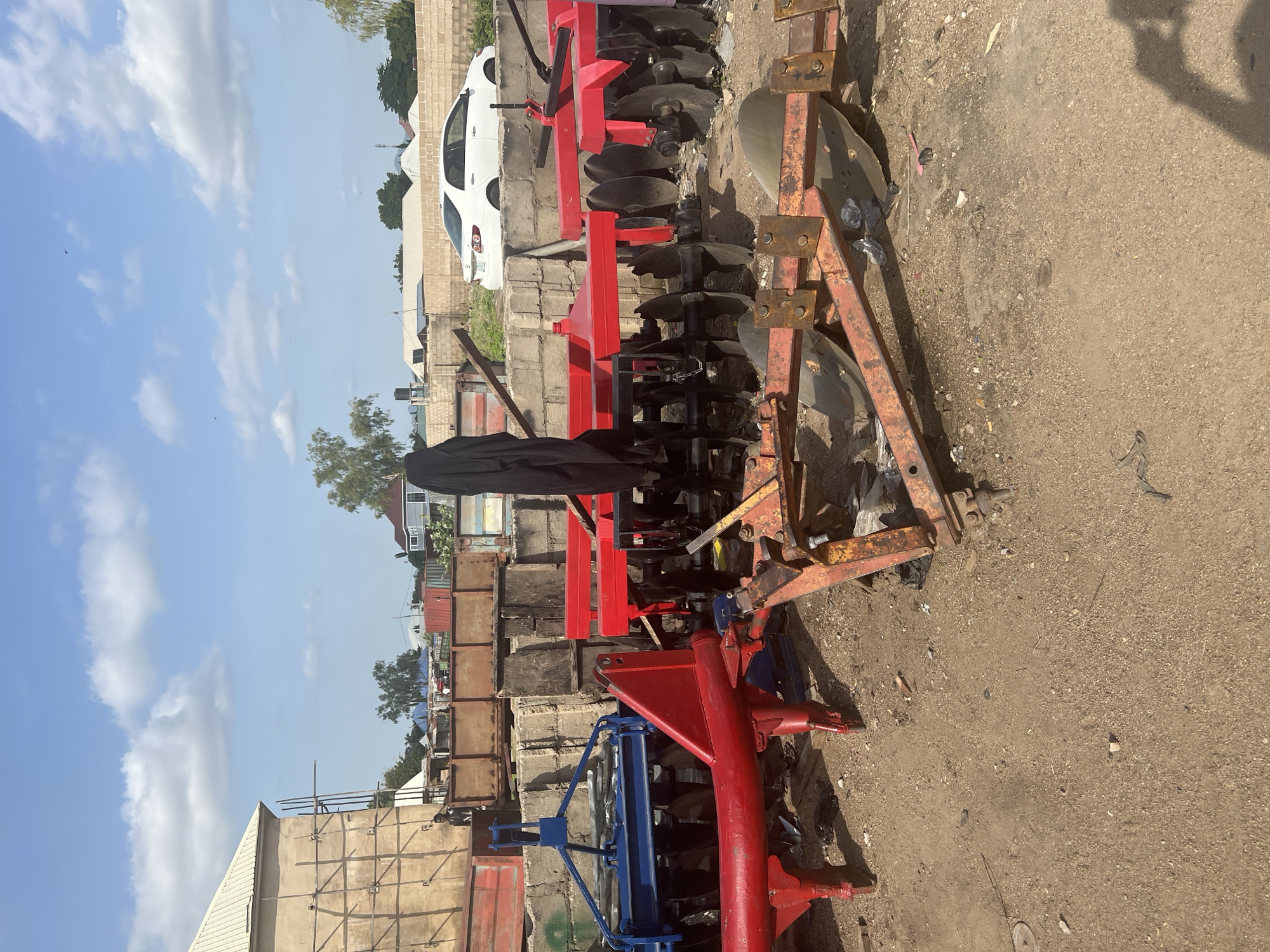 The image depicts an outdoor storage area in Jalingo, Nigeria, filled with agricultural equipment. In the foreground, a red and blue disc harrow is visible, with its metal discs partially buried in the dirt. To the right, a rusty orange plow with several discs is also present. Behind these, a red disc harrow with black discs is positioned. The background shows a beige building under construction with scaffolding, a white sedan, and a brick wall. The ground is covered in sand and debris. The sky is blue with scattered white clouds. There is no visible activity or human interaction in the scene.