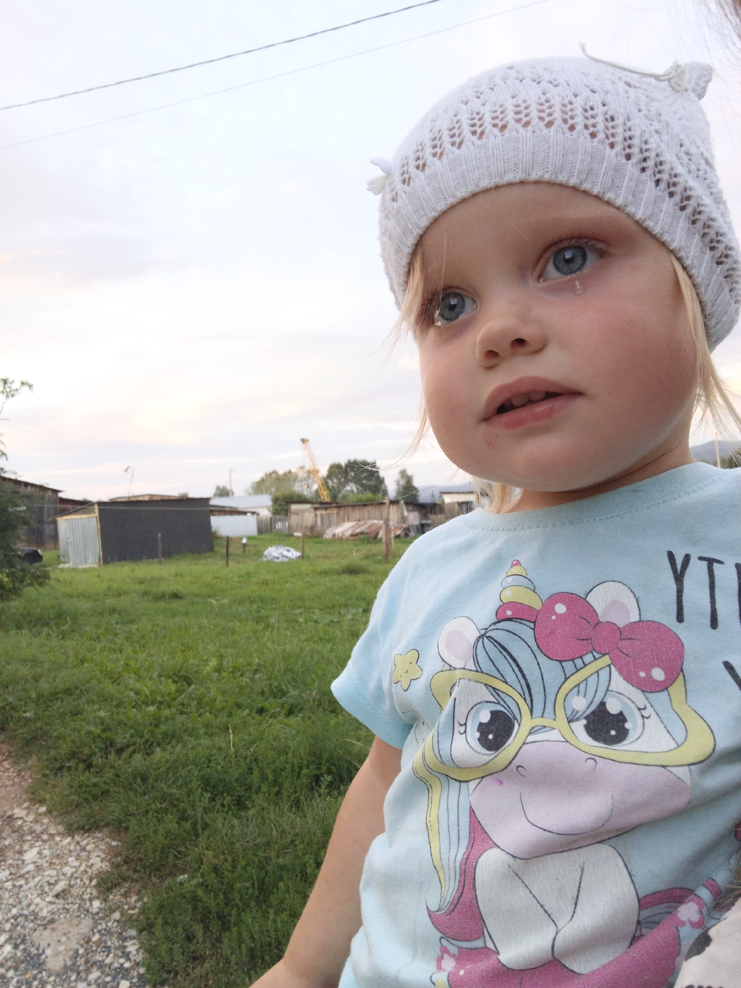 A close-up, eye-level, outdoor medium shot features a young Caucasian girl with blonde hair and blue eyes, wearing a white knitted hat with decorative bows and a light blue t-shirt adorned with a cartoon unicorn graphic. She is looking upwards and to the right, with tears visible in her eyes. Her mouth is slightly open. The background is blurred, showing a grassy field with several buildings and a yellow crane visible in the distance. The scene appears to be in Medvedyovka, Russia, based on the provided context. The lighting suggests it is late afternoon or early evening.