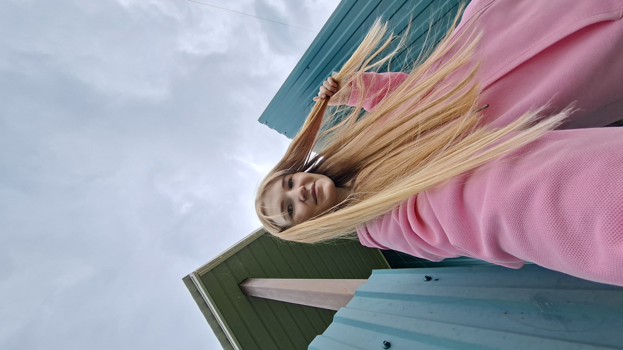 This is a medium shot, low-angle photograph of a young Caucasian woman with long blonde hair, taken from a ground-level perspective looking up. The woman is wearing a pink hooded sweatshirt. She is holding a section of her hair in her right hand, allowing it to cascade down around her. Her expression is gentle and she appears to be smiling slightly, with her eyes looking towards the camera.
The background consists of a cloudy, overcast sky and portions of a green and teal corrugated metal structure, likely a building or shed, with a wooden beam visible. The composition is dynamic due to the low angle and the way the woman's hair is blown around, suggesting a breezy outdoor setting. The time of day is indeterminate due to the overcast sky, but the lighting suggests it is daytime. The setting appears to be outdoors, possibly in a rural or semi-rural area, given the building materials and open sky. There is no visible text in the image.