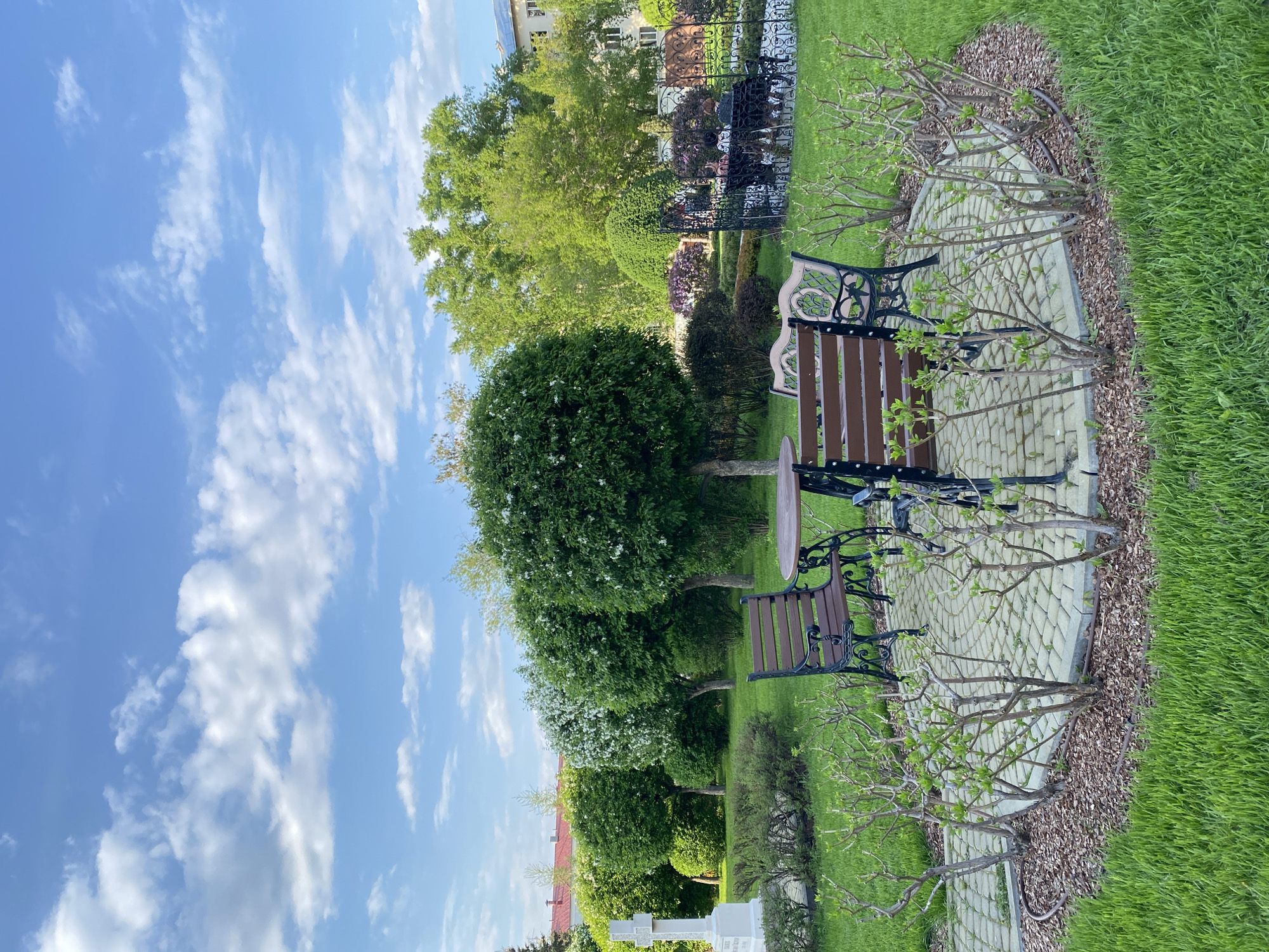 The image shows a well-maintained park or garden on a sunny day. The sky is blue with scattered white clouds. Two benches with a small round table stand on a paved walkway made of light-colored bricks. Small trees or bushes, some with white blossoms, line the pathway. Beyond the pathway is a stretch of green grass. In the background, various trees and shrubs are visible, along with sections of a black wrought-iron fence and glimpses of buildings. The location is likely within the city of Yekaterinburg, Russia, but specific details confirming the exact location within the city are absent from the provided image.