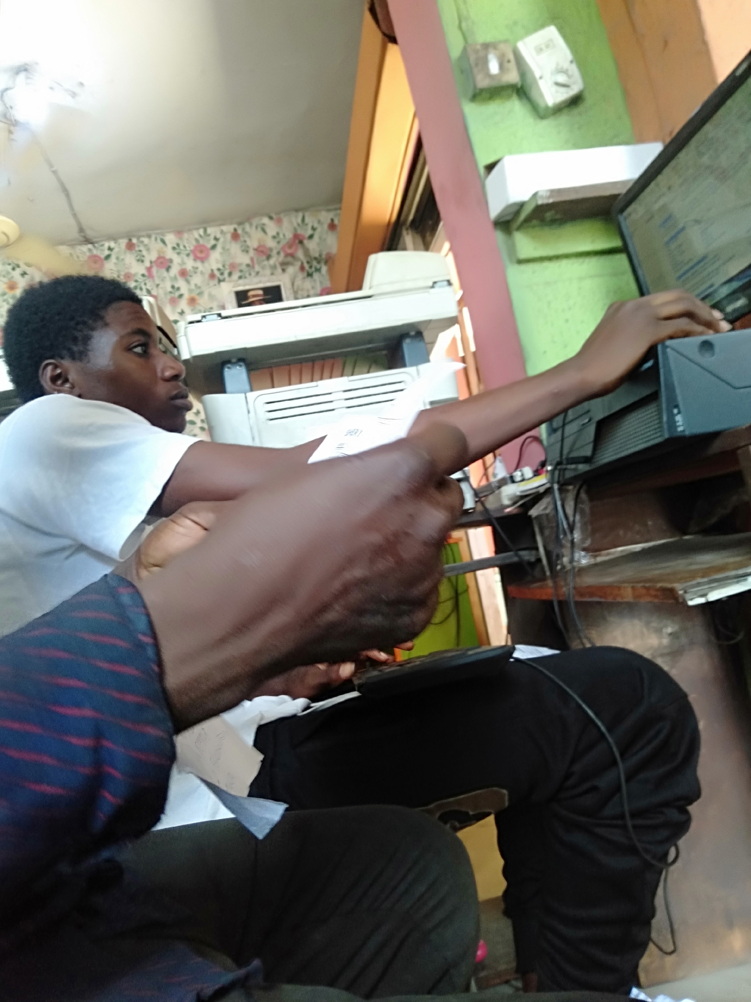 A young Black man in a white t-shirt is operating a computer. He is seated, with his right arm extended towards the computer tower, which sits on a cluttered desk. The computer screen displays text. In the foreground, another person, partially visible, has on a blue and red striped shirt. Black pants are prominent in the mid-ground. A large printer is positioned behind the computer. The setting appears to be an indoor office space with floral wallpaper and pale green walls. Two electrical boxes are visible on the wall. The specific location in Shangisha, Nigeria, cannot be determined solely from the image.