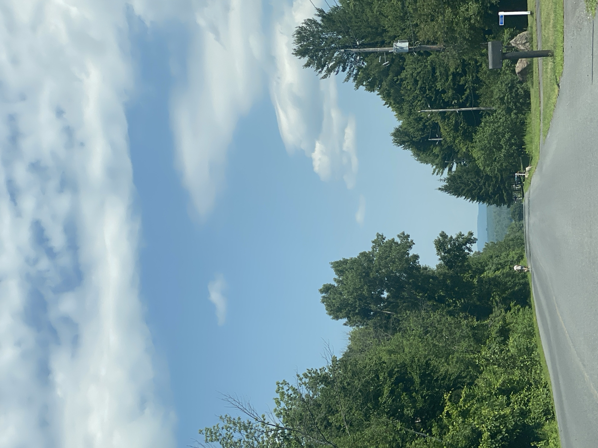 A drone shot captures a tree-lined road leading into the distance, under a bright blue sky with scattered clouds. Dense green foliage flanks both sides of the asphalt road, with a single person walking on the side of the road on the left. Further down the road, on the right, a mailbox stands next to a large rock. The landscape recedes into a hazy horizon with rolling hills. The scene appears to be a rural setting in East Hawkesbury, Canada.