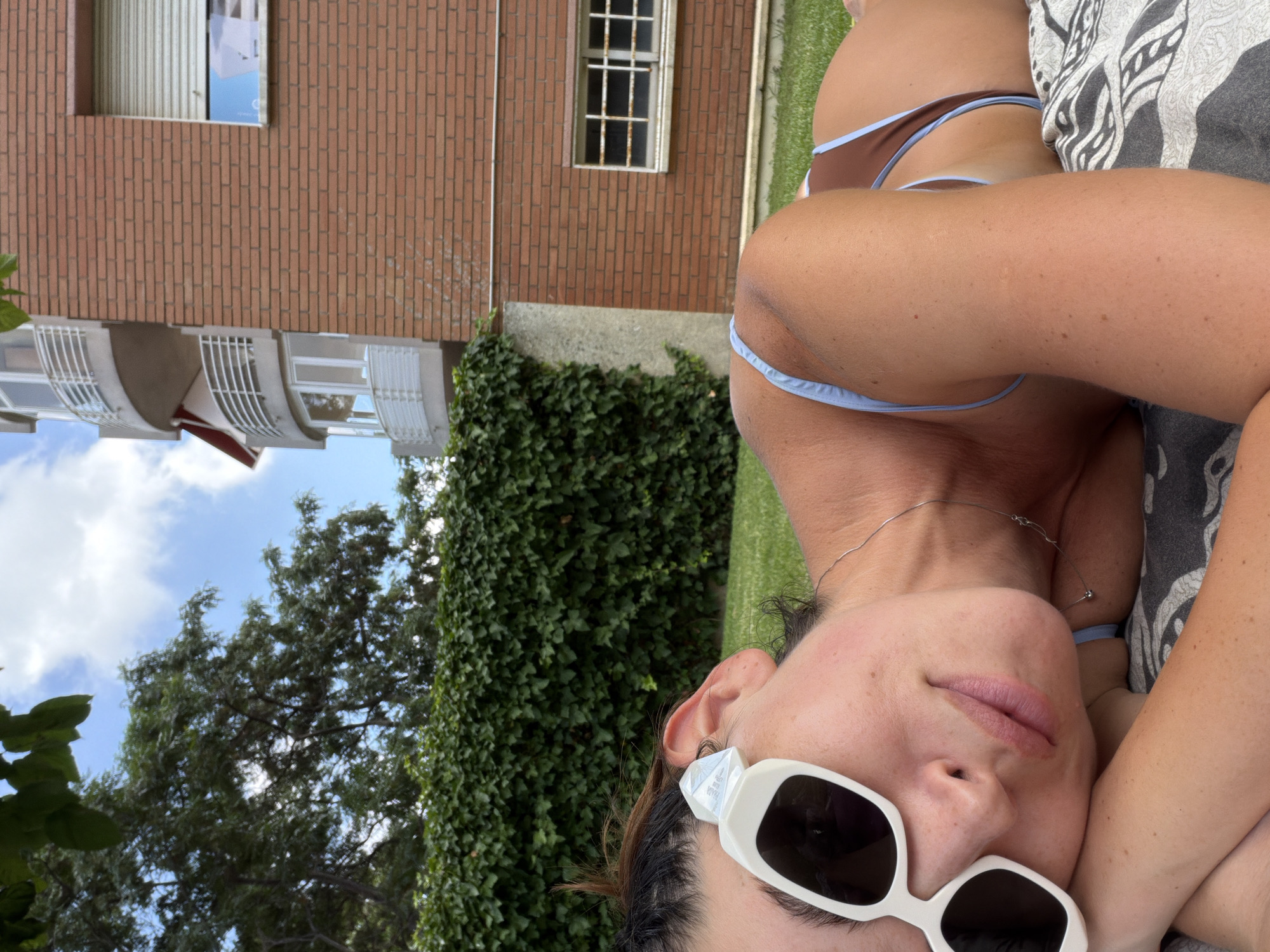A young woman with dark hair and white-rimmed sunglasses lies on her side on a patterned blanket on a grassy lawn. She wears a brown bikini with light blue trim. Her left arm is bent, supporting her head. Behind her, a dense green hedge forms a backdrop, with a brick building and a clear blue sky with white clouds visible beyond. The building has multiple balconies with white railings and a window with a closed beige shutter. The scene appears to be a residential area, possibly a garden or patio.