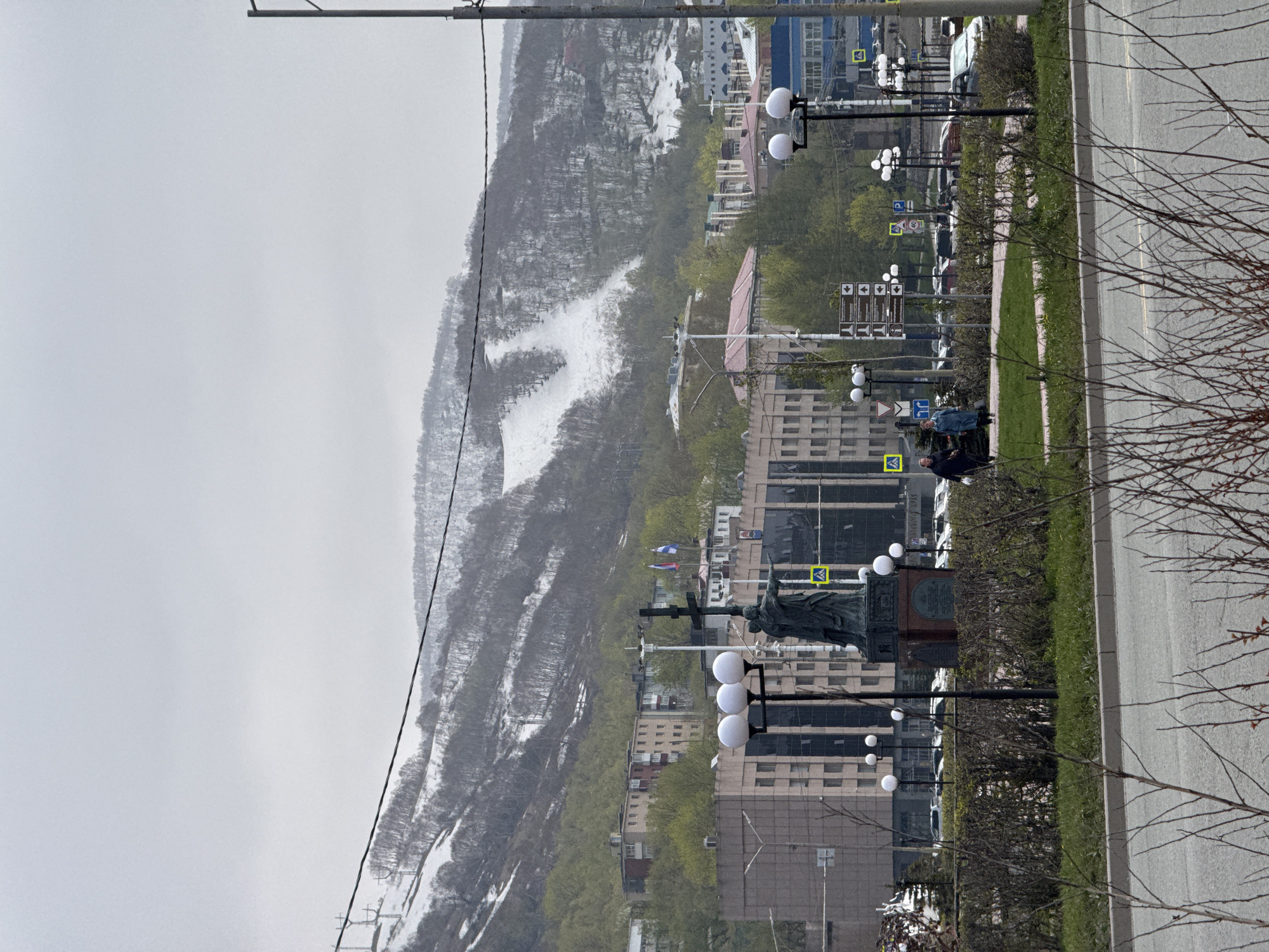 The image presents an urban landscape set against a dramatic, snow-dusted mountain backdrop, likely Petropavlovsk-Kamchatsky, Russia. The view is taken from street level, looking towards the mountain.
In the foreground and midground, a paved road runs along the right side, bordered by a grassy median and a sidewalk. Bare tree branches are visible on the far right. Along the sidewalk, numerous streetlights with spherical white lamps illuminate the area, and parked cars line the street.
A prominent feature is a dark-colored statue on a stone pedestal, situated on the sidewalk in the midground. The statue depicts a robed figure, possibly a historical personality, with one arm raised. Near the statue, two flags are visible: one with blue and white horizontal stripes, and another, possibly the Russian tricolor, displaying muted vertical stripes. Two people are standing on the sidewalk near the statue, one in a dark jacket and another in blue.
The background is dominated by a large mountain, whose slopes are covered with dark, bare trees and significant patches of white snow, suggesting either late winter or early spring. The sky is overcast, providing soft, diffuse lighting, indicating daytime. The city's buildings range from multi-story structures with modern glass facades to older, less distinct buildings.
Visible text includes various road signs: directional arrows, parking signs ("P"), and yellow square signs with black symbols, possibly indicating pedestrian crossings. An inscription plate is visible on the statue's pedestal, but its text is illegible from this distance. The overall scene captures a city coexisting with a rugged, snow-covered natural environment during a transitional season.