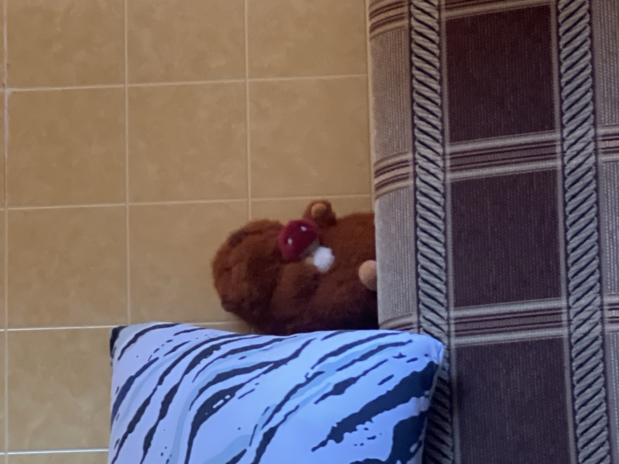 A brown stuffed gnome with a red mushroom hat and white accents is sitting on the armrest of a sofa. To the left of the gnome is a large pillow with a black and white stripe pattern. The background consists of tiled walls. The scene is indoors, likely in a residential setting.