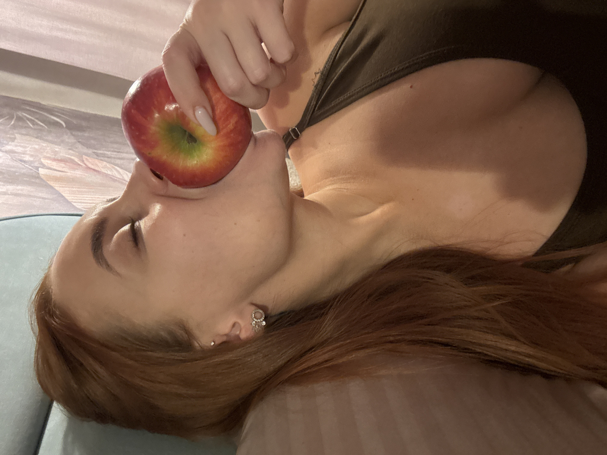 A woman lies on a light blue surface, presumably a bed or couch, in what appears to be an indoor setting. She is holding a red apple against her face with her right hand. She has reddish-brown hair, fair skin, and is wearing an earring in her left ear. She is wearing a dark green top. The background includes a floral patterned surface. The location is unknown and not discernible from the image.